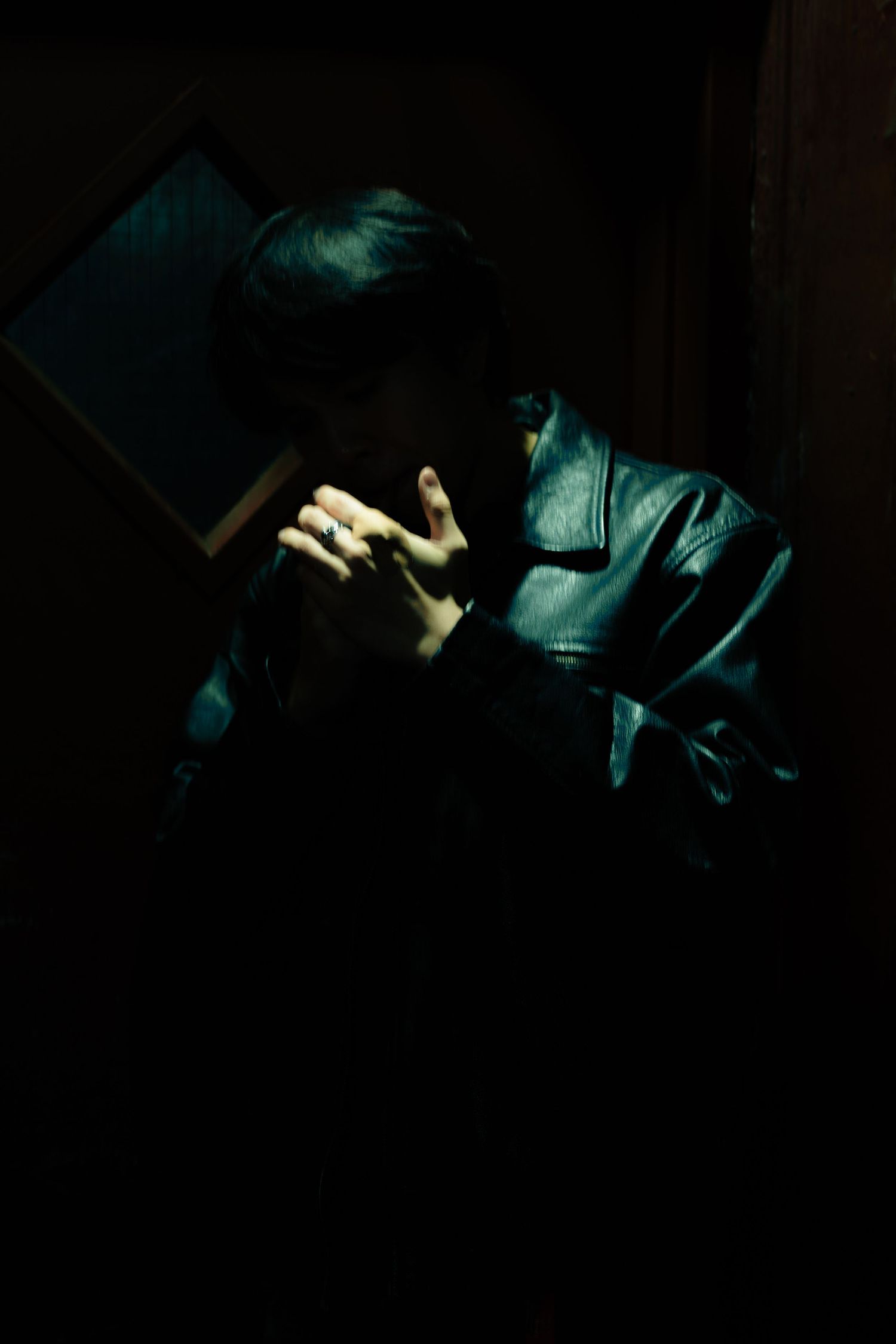 Person in a black leather jacket sits in dramatic low-key lighting, hand raised near the face, with a framed window in a dark room.