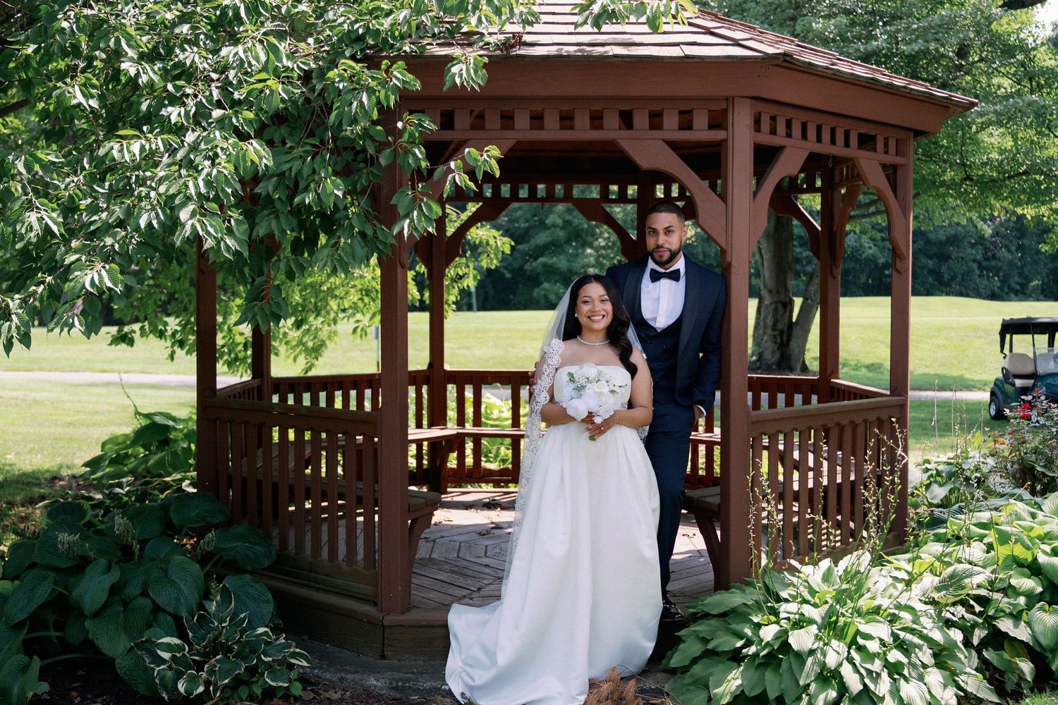 Bride and groom posing under a wooden gazebo surrounded by lush greenery on their wedding day.