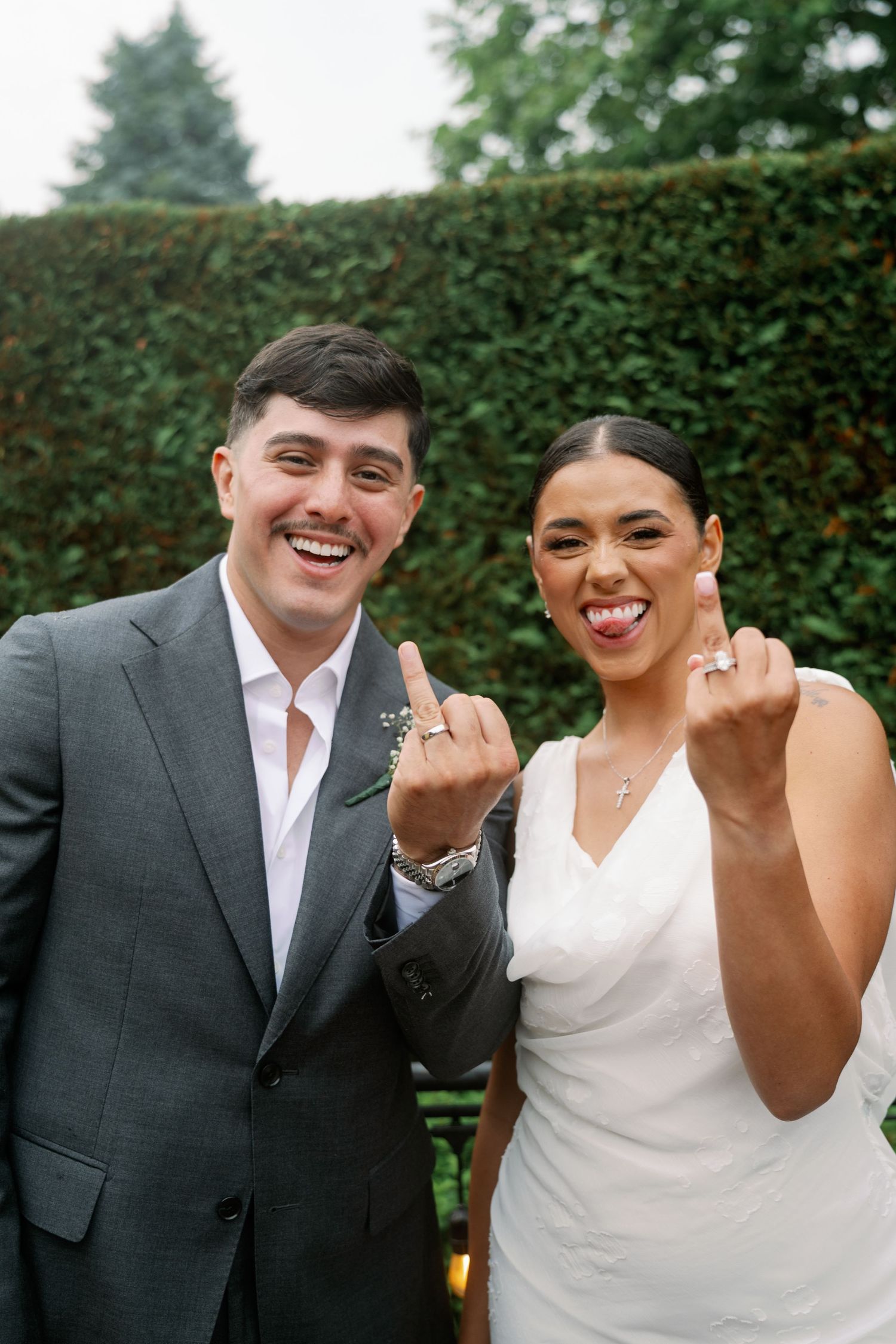 Happy newlyweds showing off wedding rings, bride in white dress and groom in gray suit, posing outdoors near green hedge.
