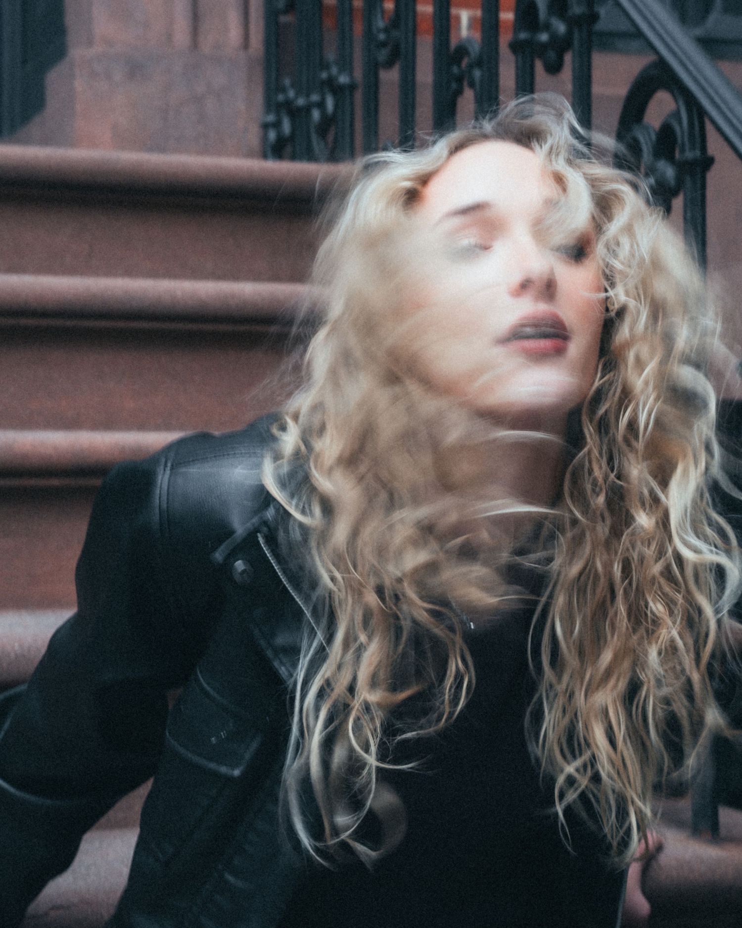 Woman with long, wavy blonde hair in a black leather jacket tosses her head in motion blur on a brownstone stoop with iron railings.