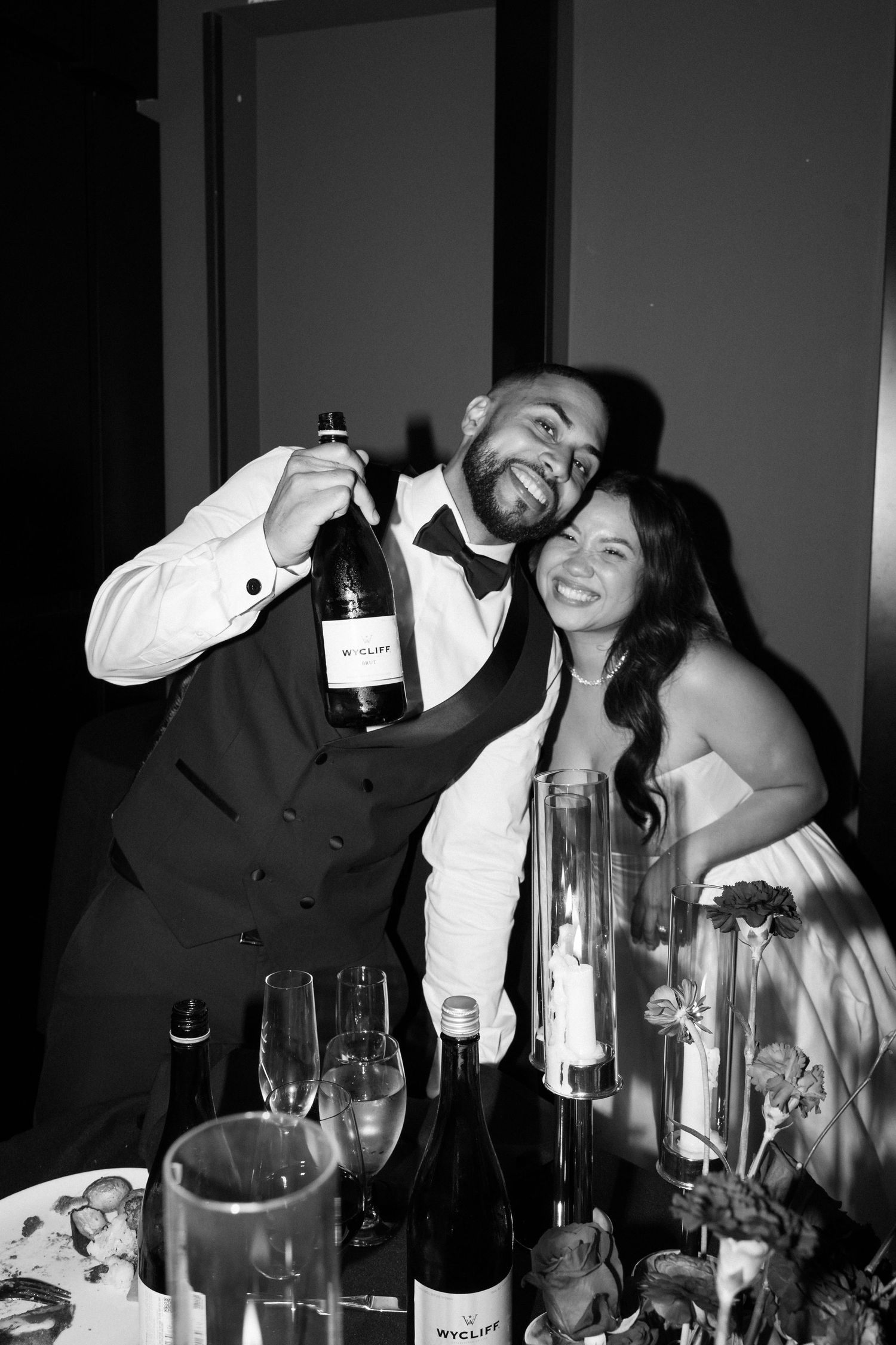 Black and white photo of a smiling couple at a wedding reception table, man holding a wine bottle.