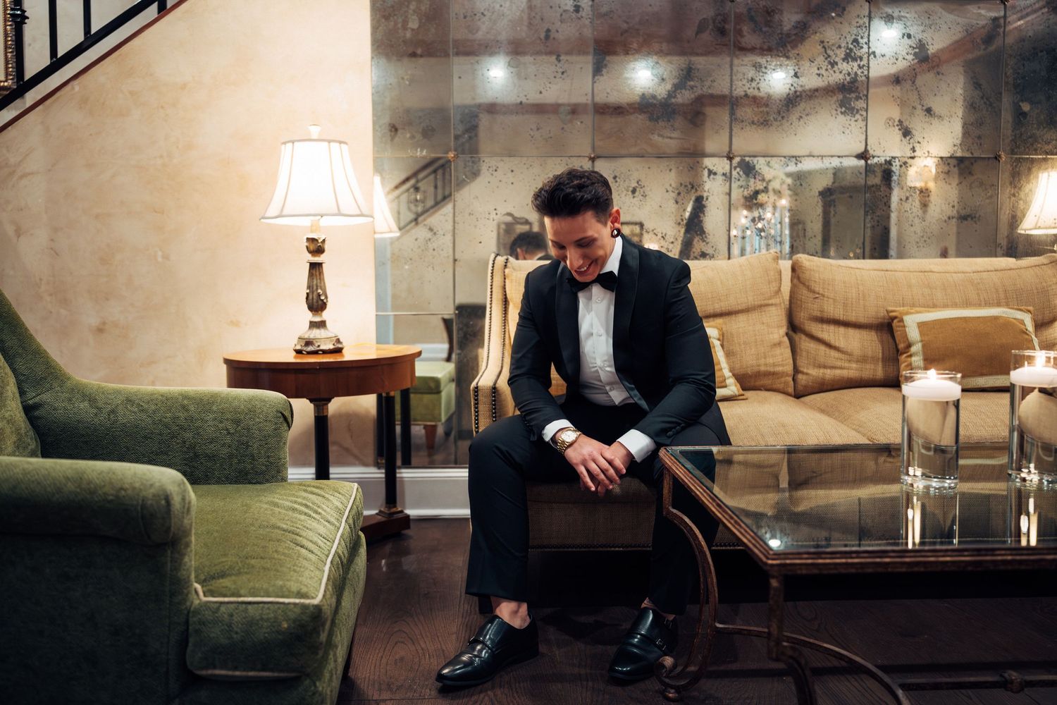 A well-dressed man in a black tuxedo and bow tie sits pensively on a bench in an elegant vintage lounge.