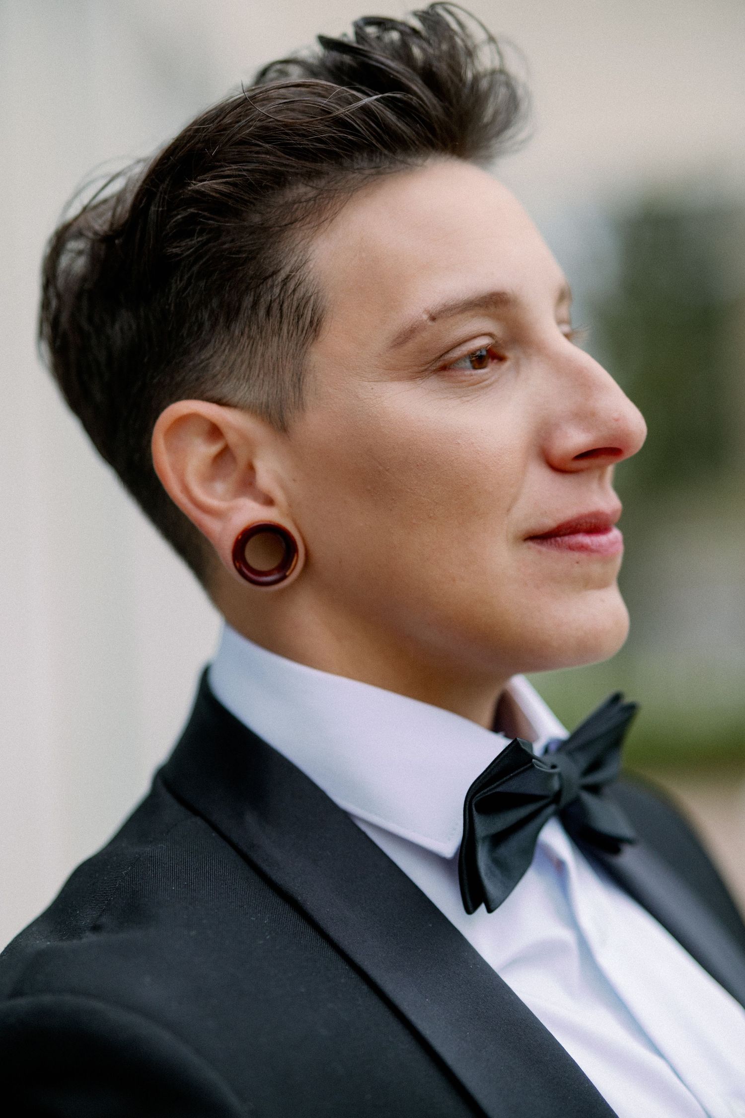 Person in tuxedo with bow tie and ear gauges, short undercut hair, profile view, formal attire.