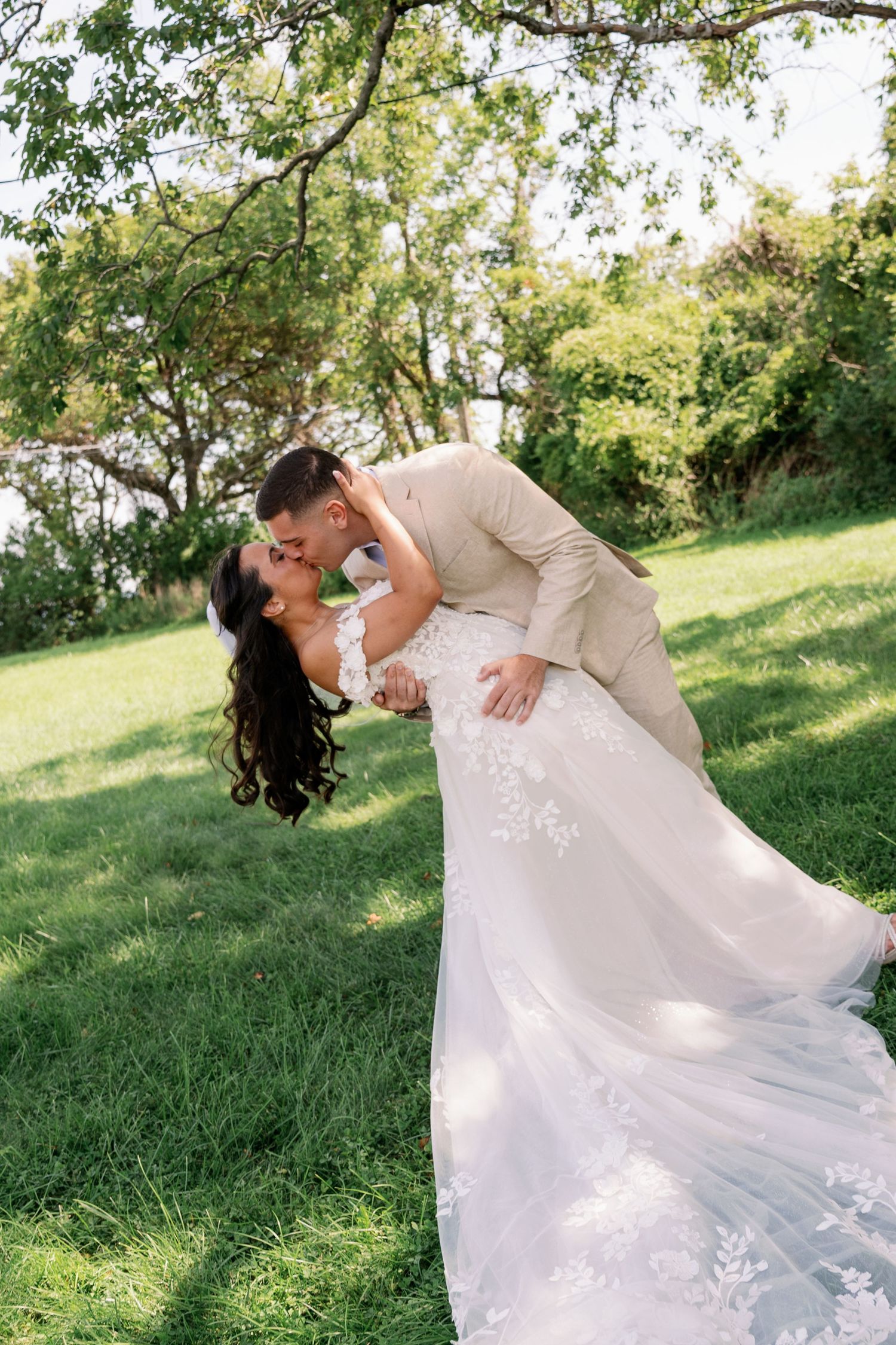 Groom in a tan suit dips and kisses his bride in a lace wedding gown on a sunny green lawn surrounded by trees.