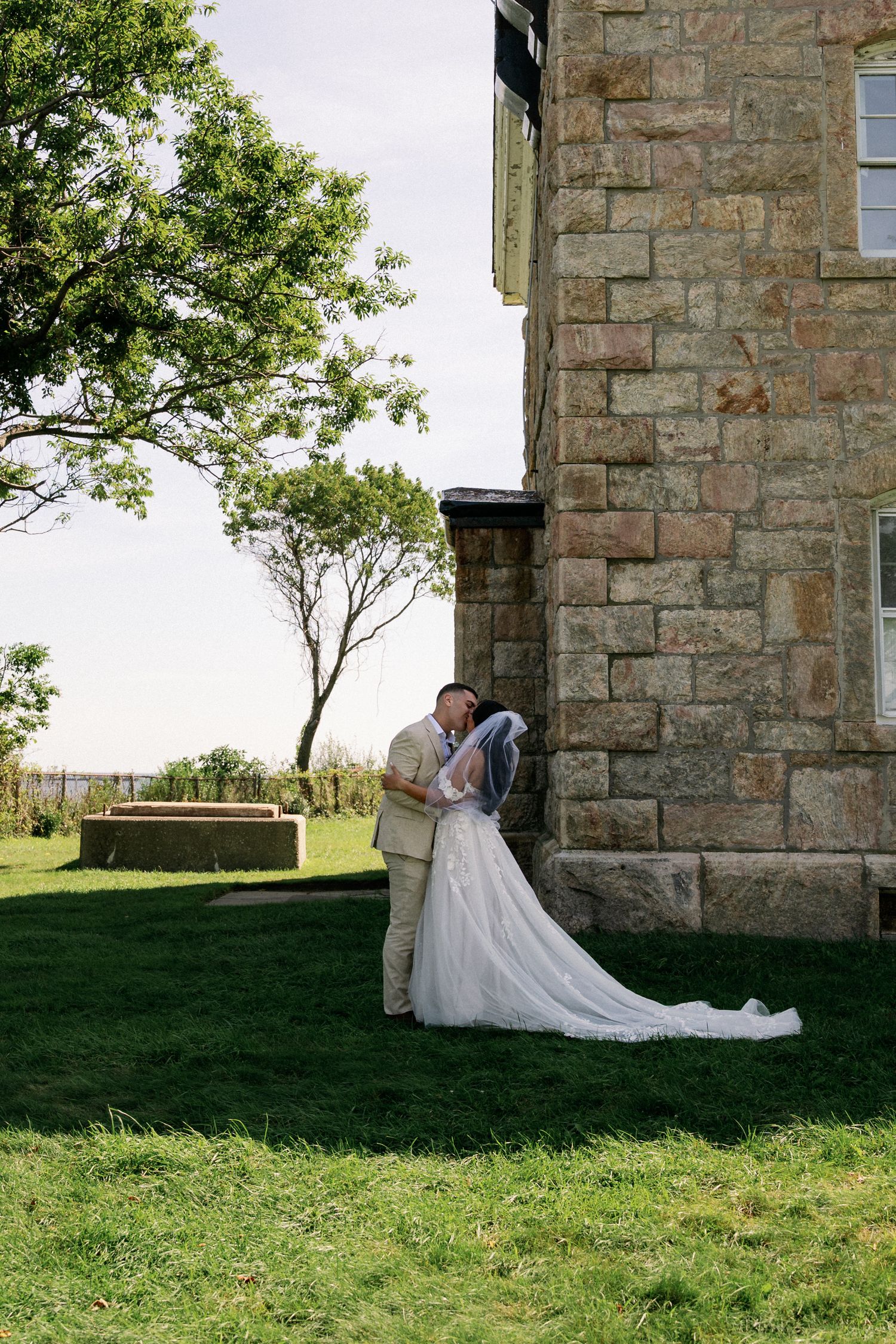 Bride in a white gown and veil embraces her groom beside a historic stone building on a sunny, grassy lawn.