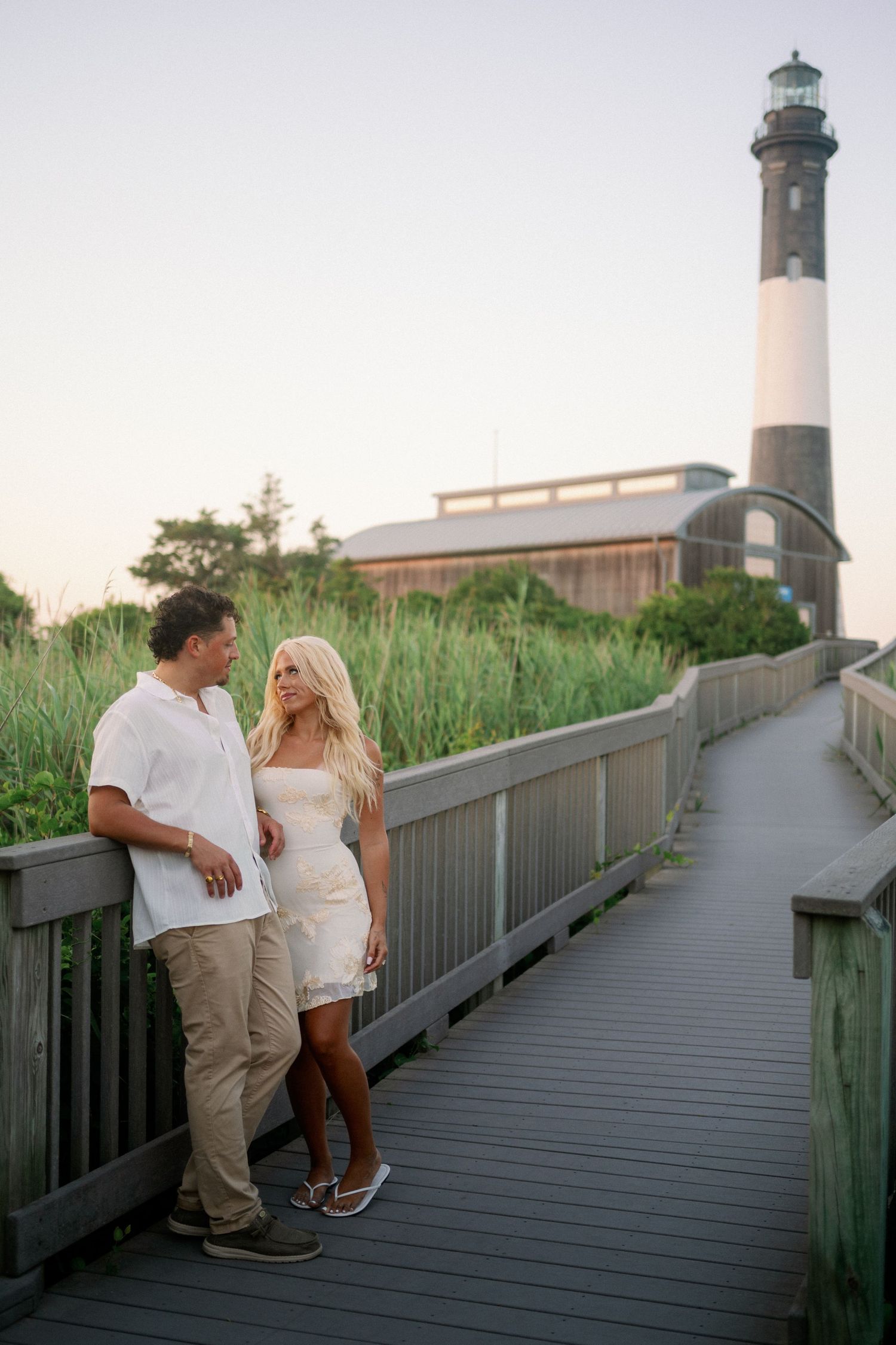 Couple in white outfits on a wooden boardwalk near a lighthouse surrounded by lush greenery at sunset.