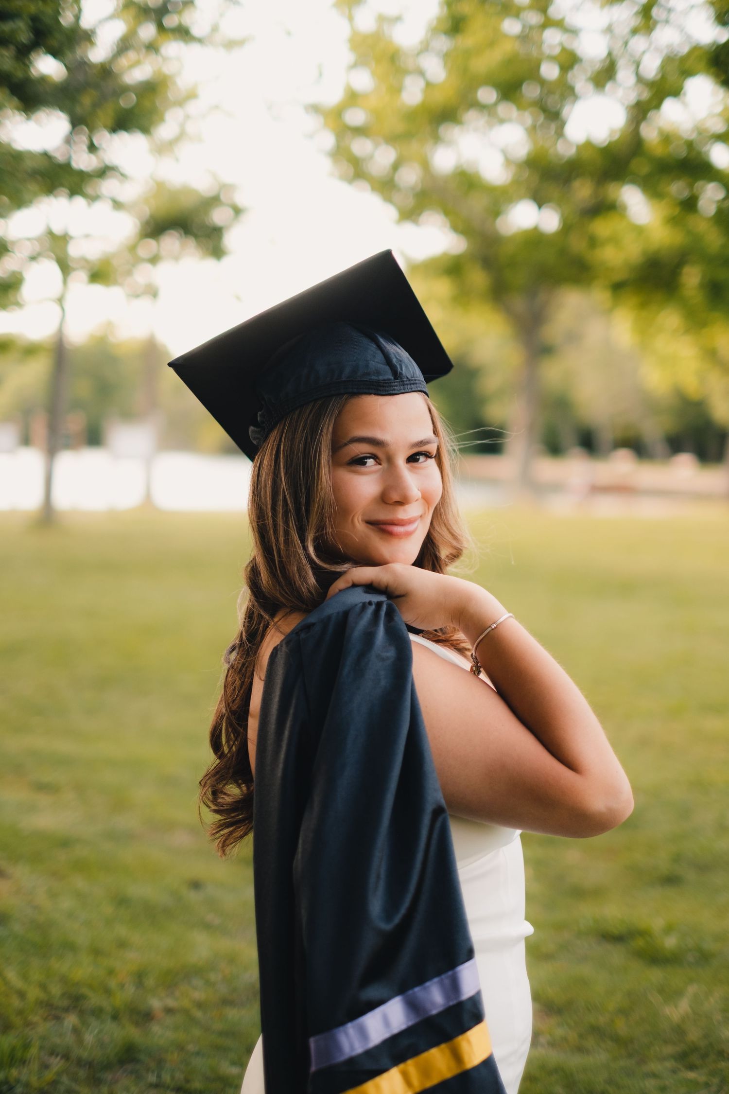 Graduate posing outdoors in a cap and gown against a sunny green background.