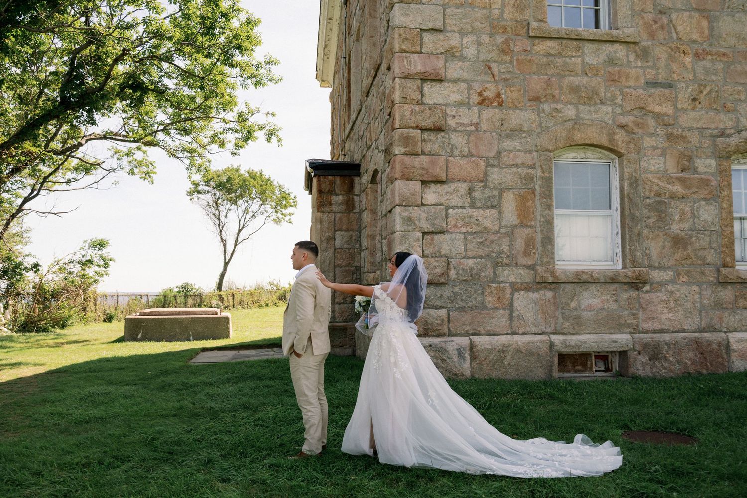 Bride in a white gown and veil reaches toward the groom beside a historic stone building on a sunny grassy lawn at a wedding.