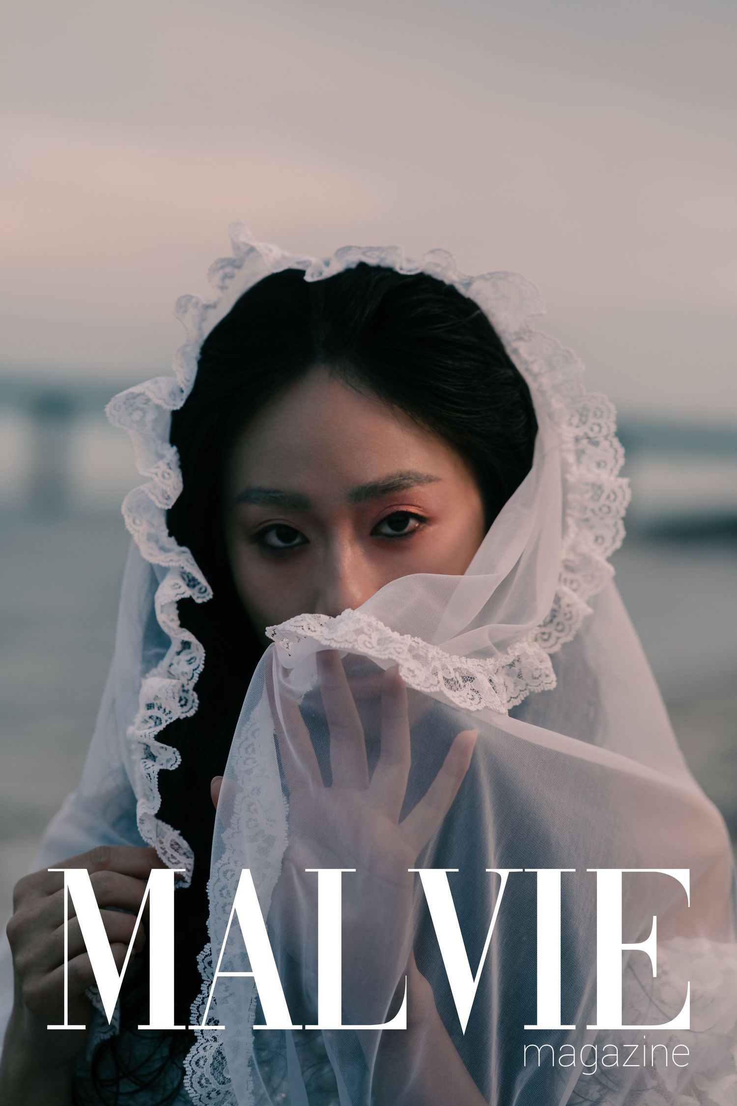 Woman in white lace veil cover on MALVIE Magazine, elegant editorial fashion photography with moody coastal backdrop.