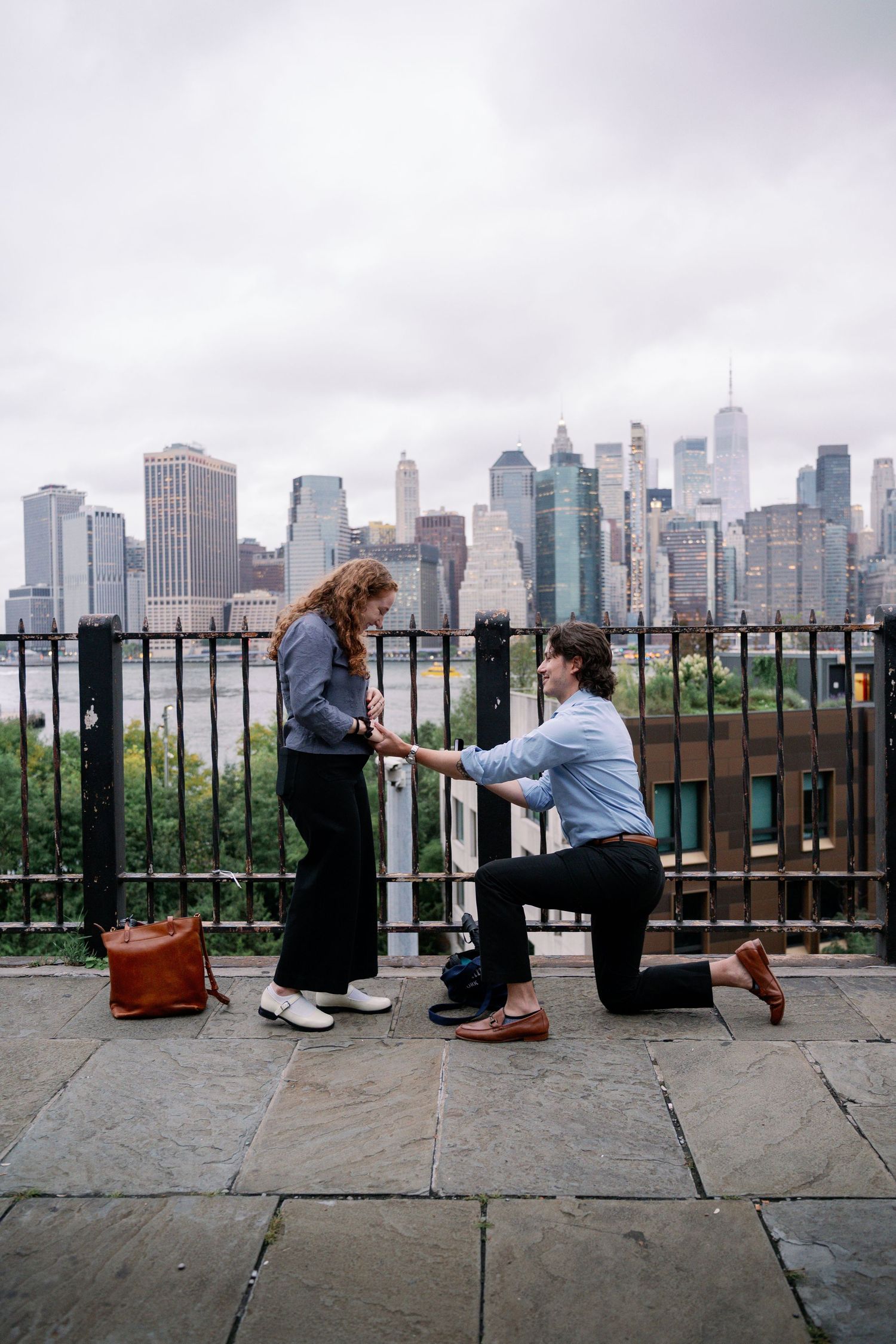 Person kneeling on one knee proposing to their partner on a stone terrace with a hazy city skyline in the background.