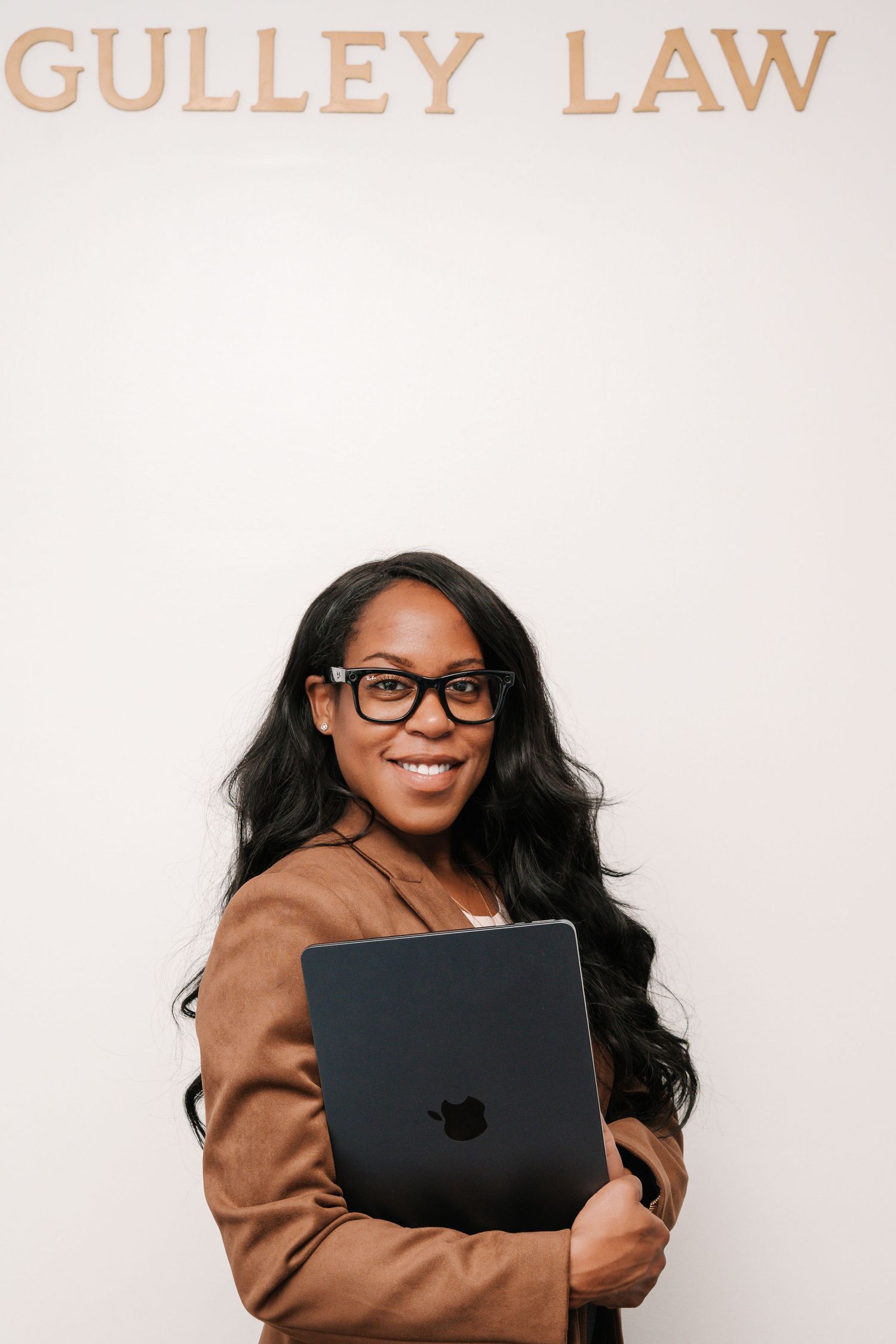 Smiling woman in glasses and a brown blazer holds a laptop in front of a wall with Gulley Law lettering.
