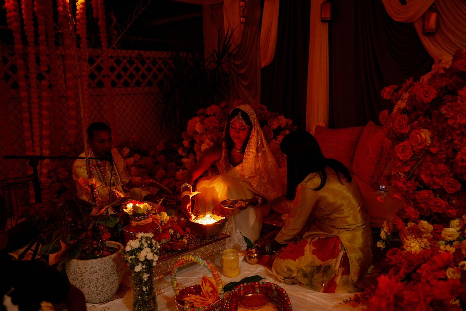 Women in traditional attire perform a candlelit ritual surrounded by red flowers and warm lighting at a festive Indian wedding ceremony.