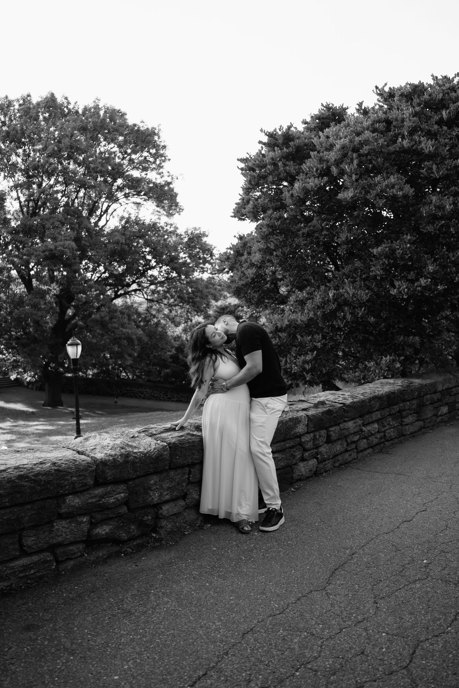 Black and white photo of a couple kissing against a stone wall in a park, surrounded by lush trees.