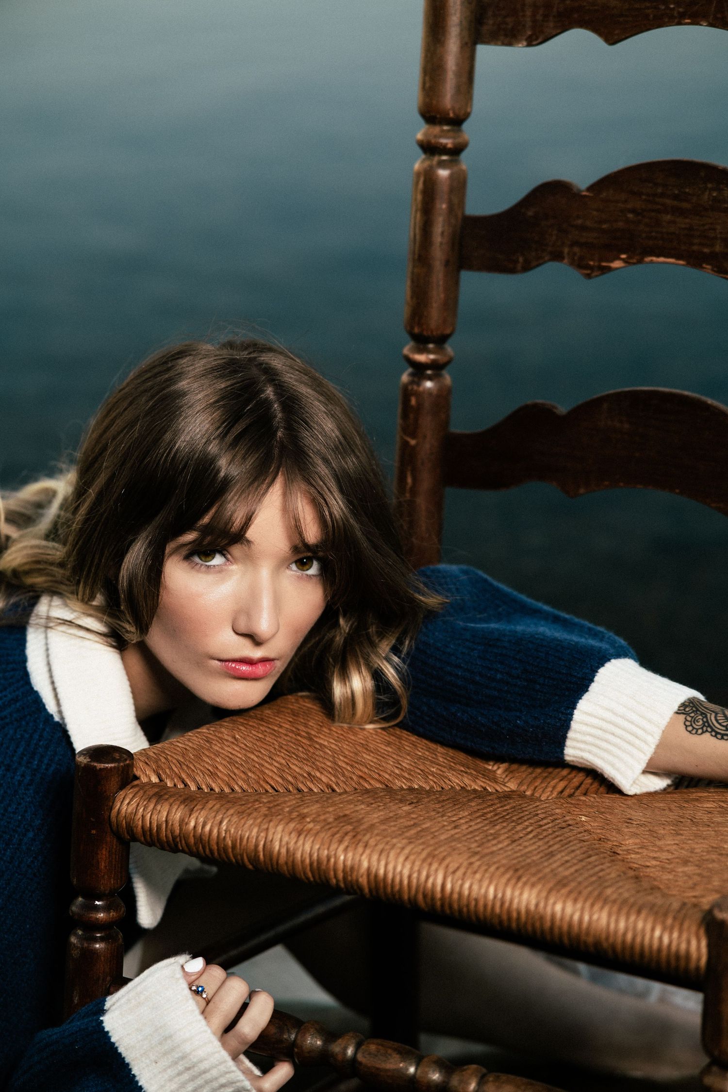 Artistic portrait with a navy sweater and white collar against a woven chair and water background.