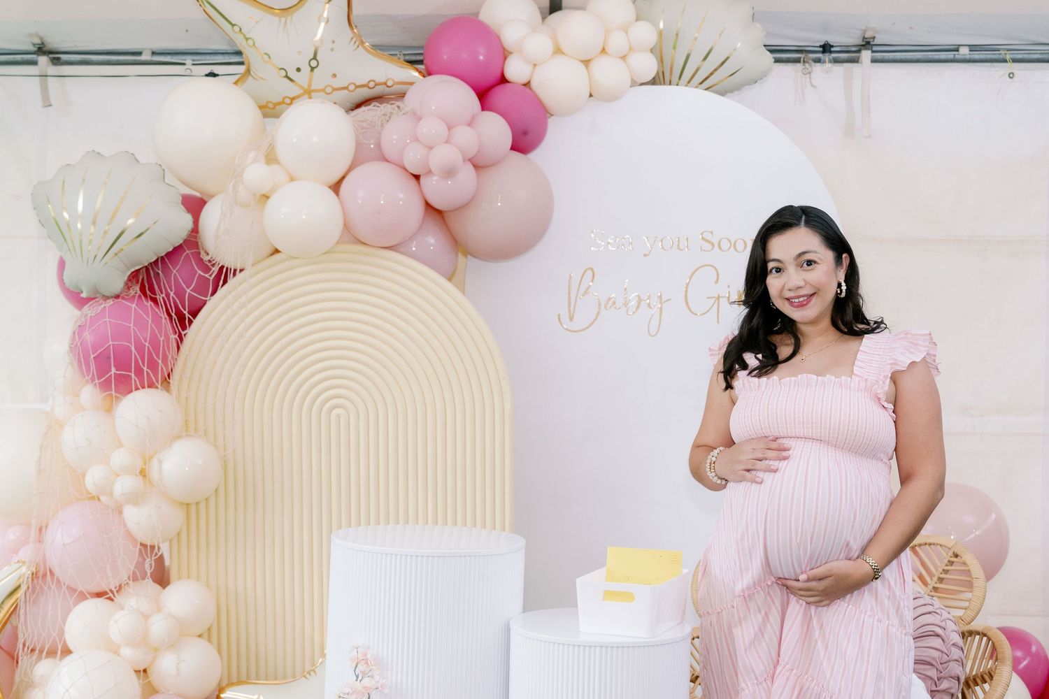 Pregnant woman in a pink ruffled dress cradles her belly at a baby shower with pastel balloons, an arch backdrop and a dessert table.