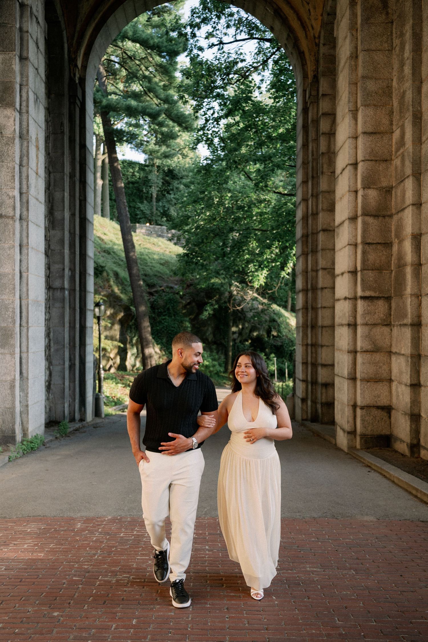 Couple walking hand-in-hand under a stone arch in a lush park, man in black shirt and woman in white dress.