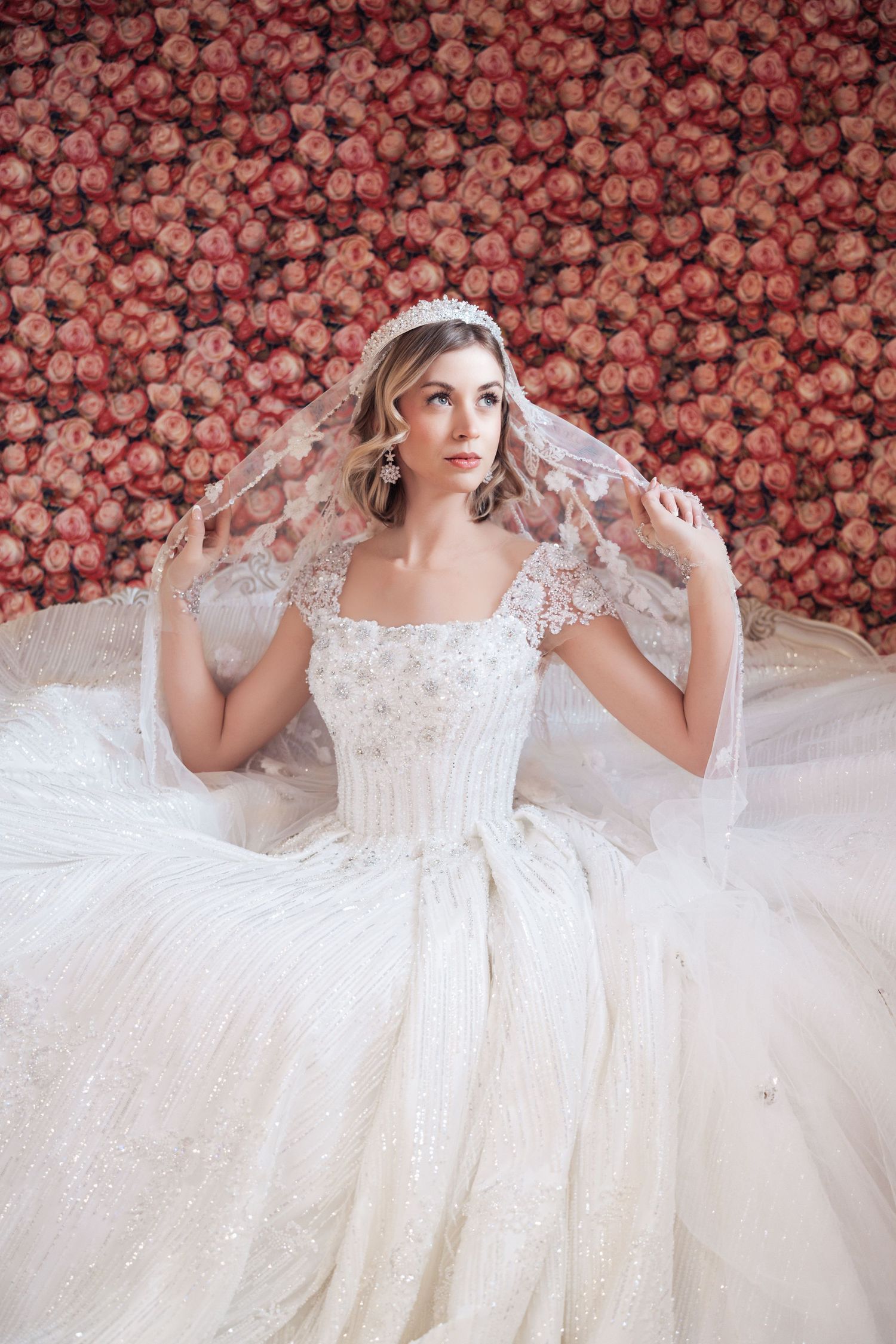 A dreamy bridal portrait against a backdrop of pink roses featuring an elegant white lace wedding gown.