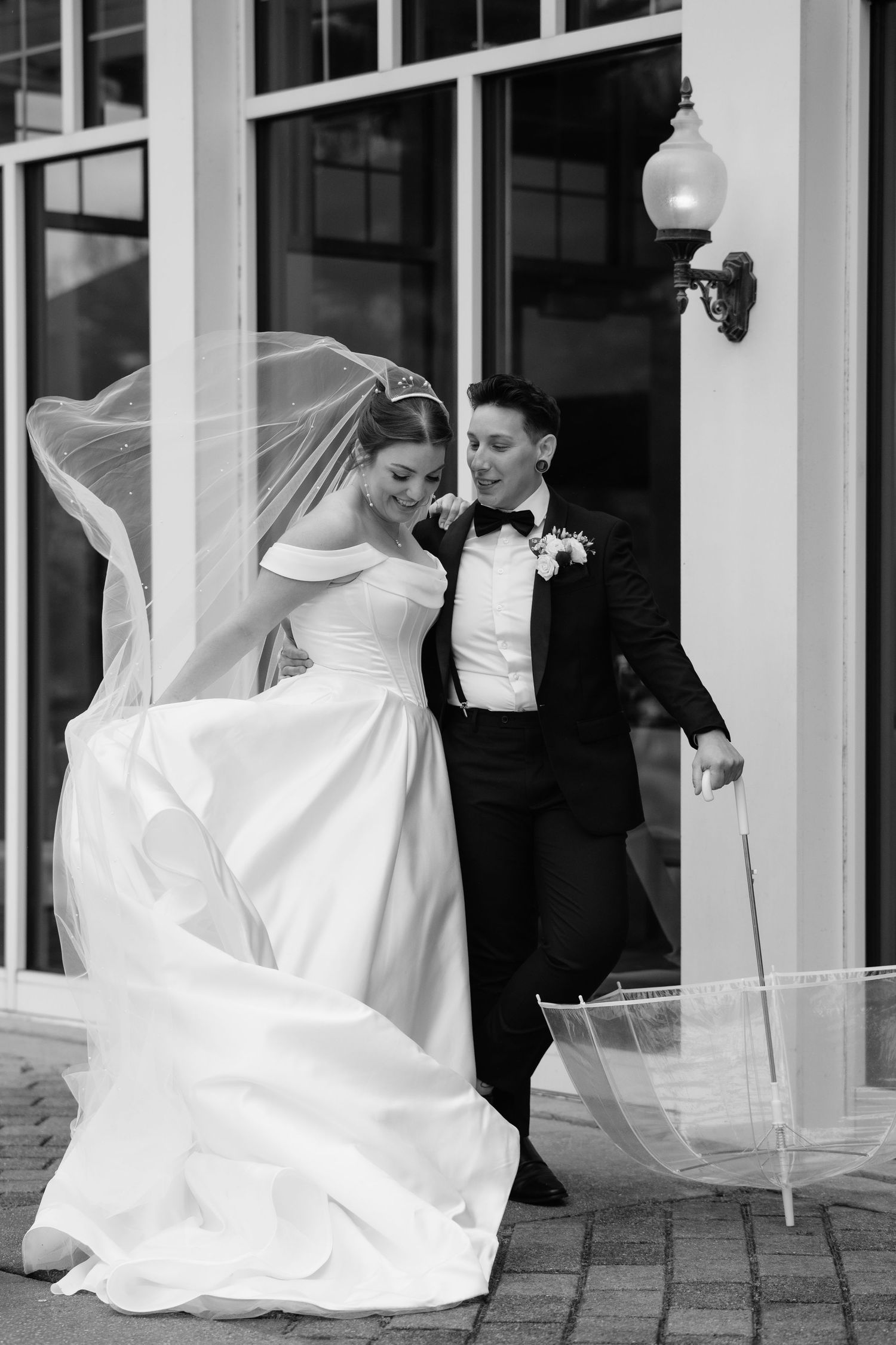Black and white photo of a bride and groom exiting a building, veil flowing in the wind, groom holding a clear umbrella.
