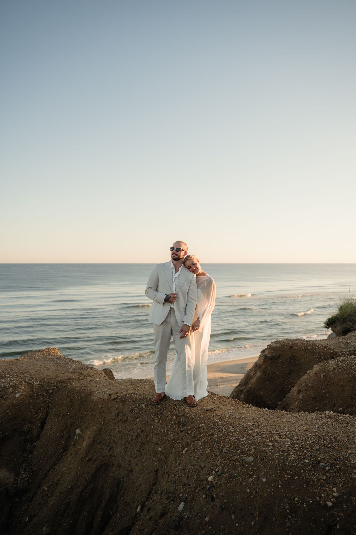 Couple in white outfits embracing on a sandy coastal cliff overlooking the ocean at sunset during a beach wedding photoshoot.