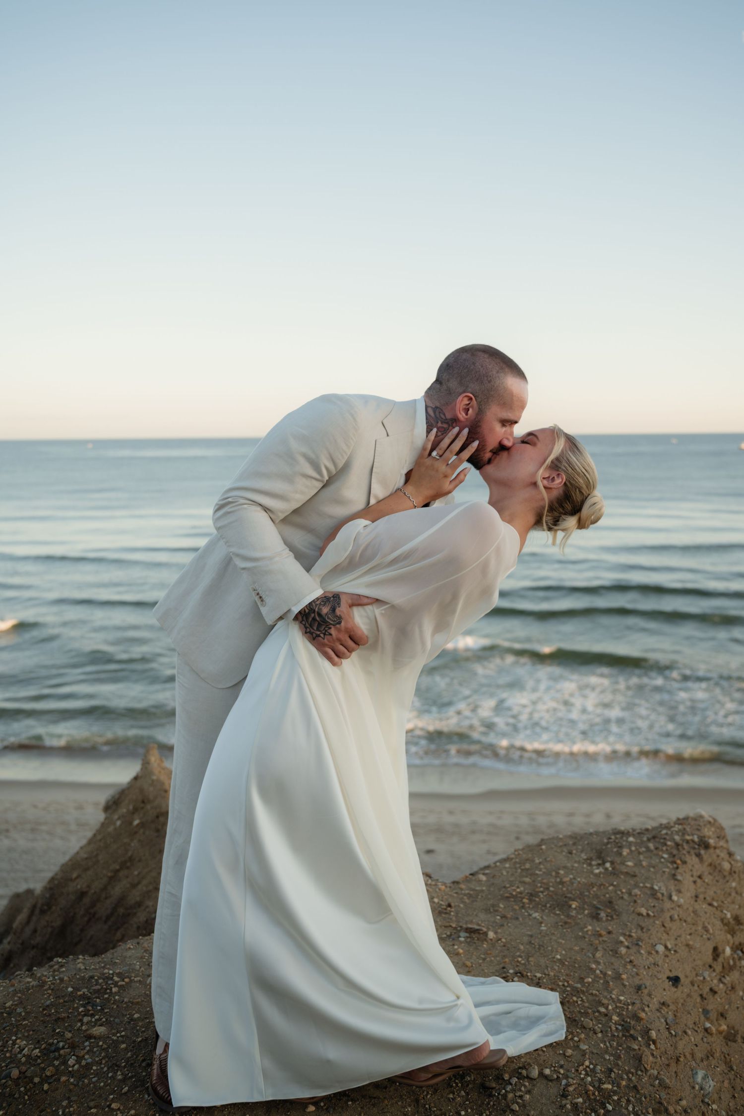 Bride in a flowing white gown and groom in a cream suit share a dipped kiss on a rocky beach at sunset with ocean waves behind them.