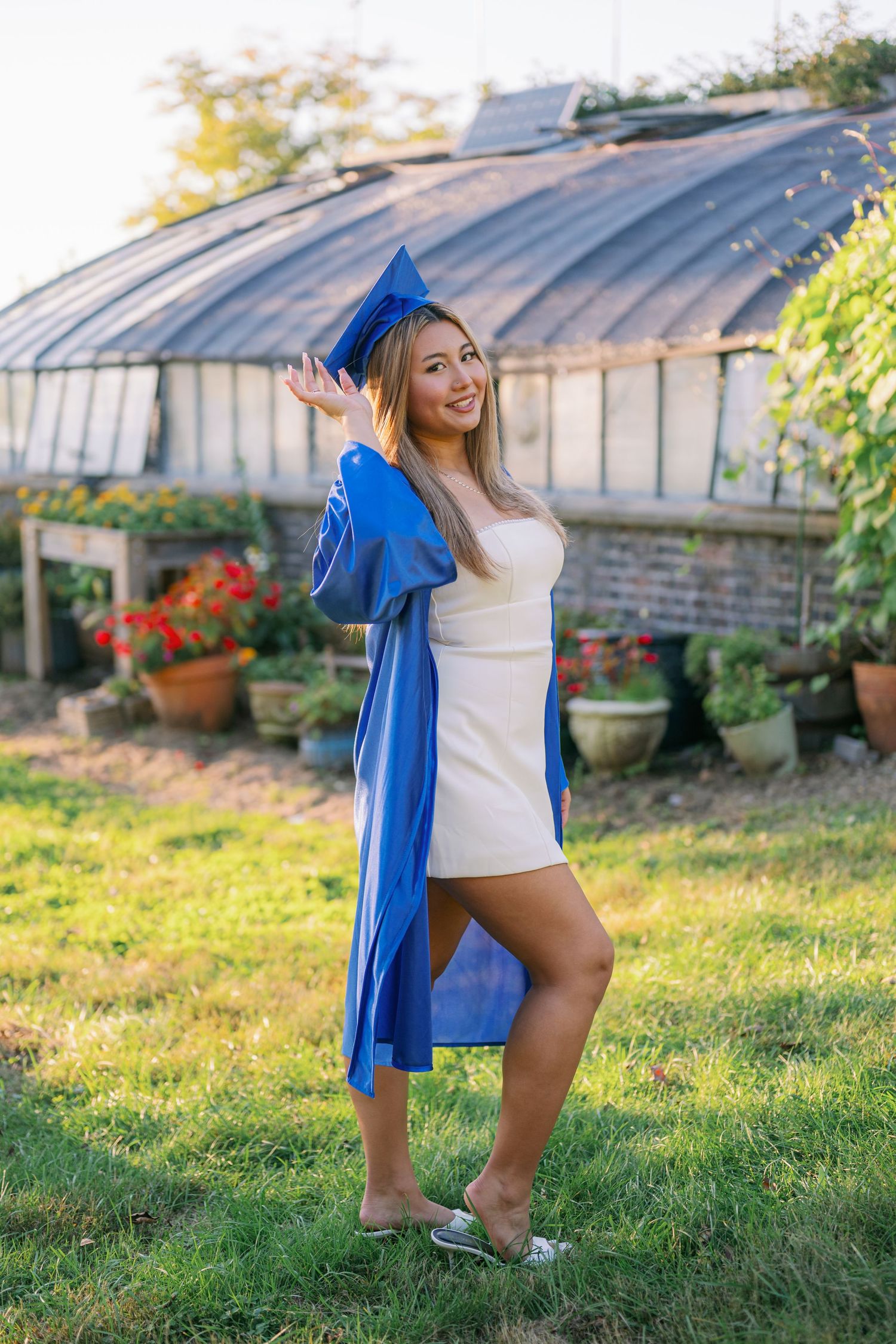 Graduate in a blue cap and gown over a white dress poses on a grassy lawn in front of a greenhouse with potted flowers.