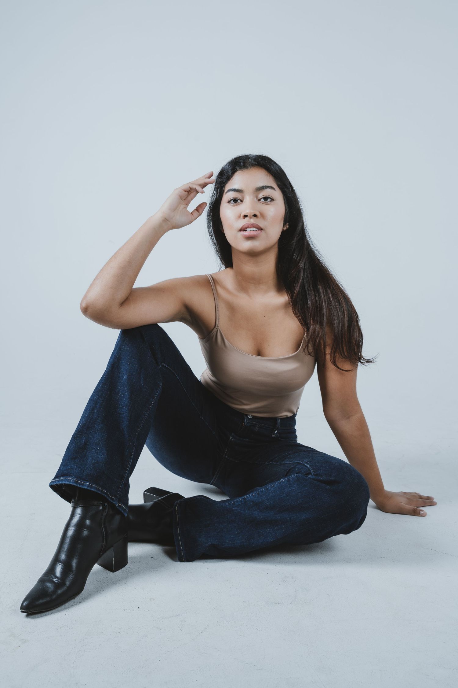 Fashion model in beige tank top, blue jeans and black boots poses sitting on white studio floor.