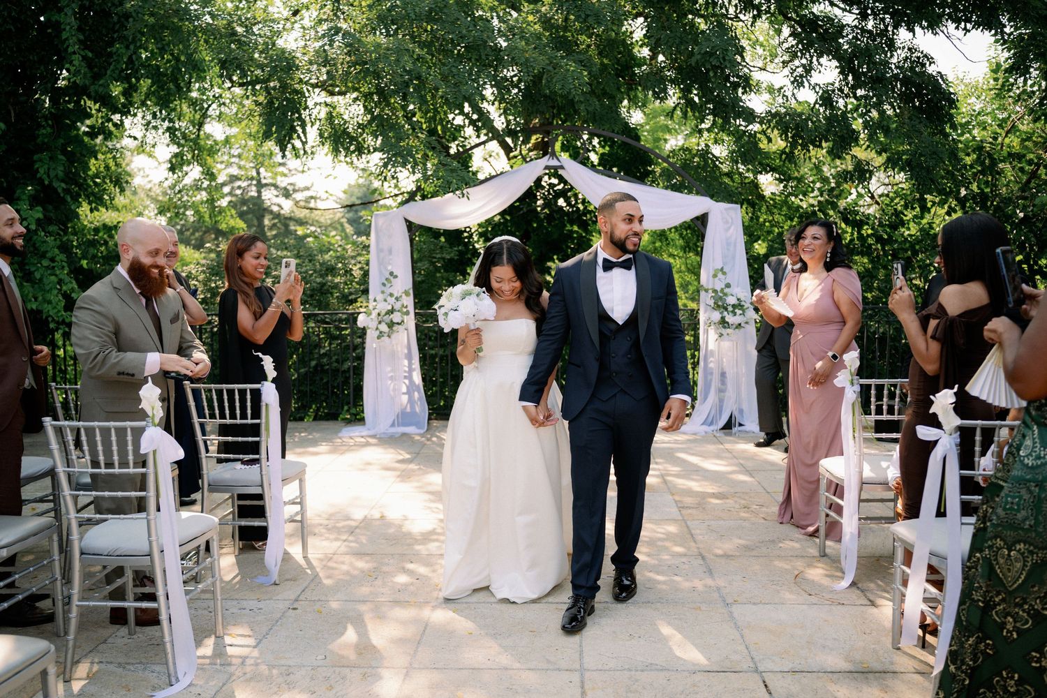 Newlywed couple walks down outdoor aisle after wedding ceremony beneath floral arch surrounded by guests.