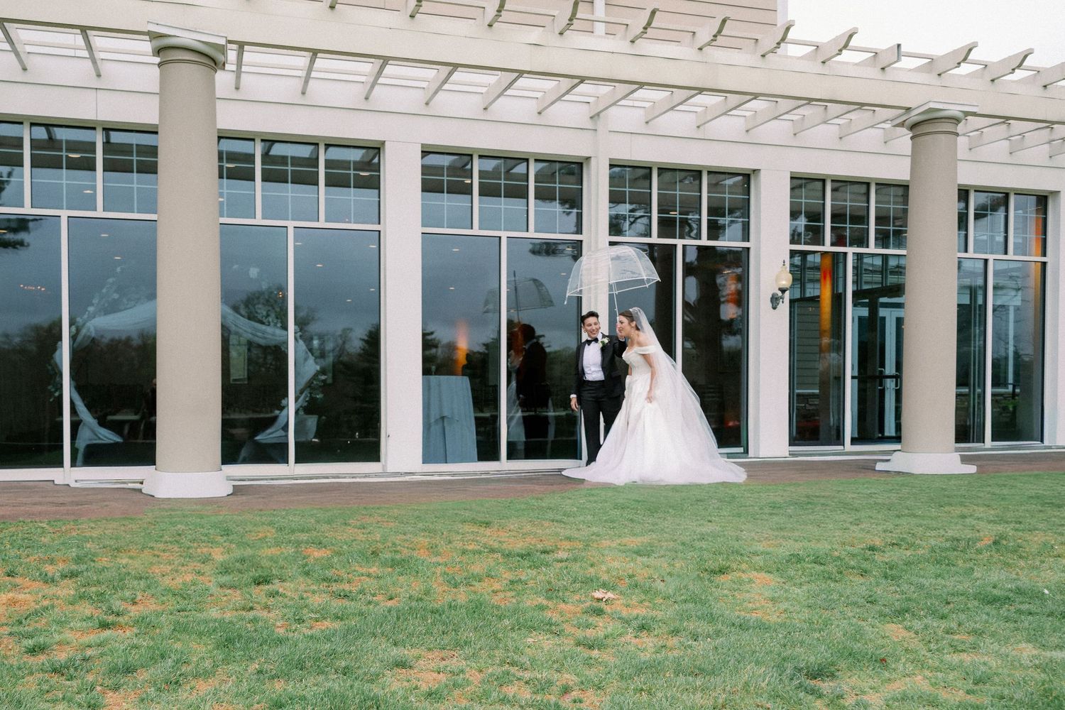 Bride and groom share a romantic moment under a clear umbrella on a lush lawn outside an elegant glass venue.