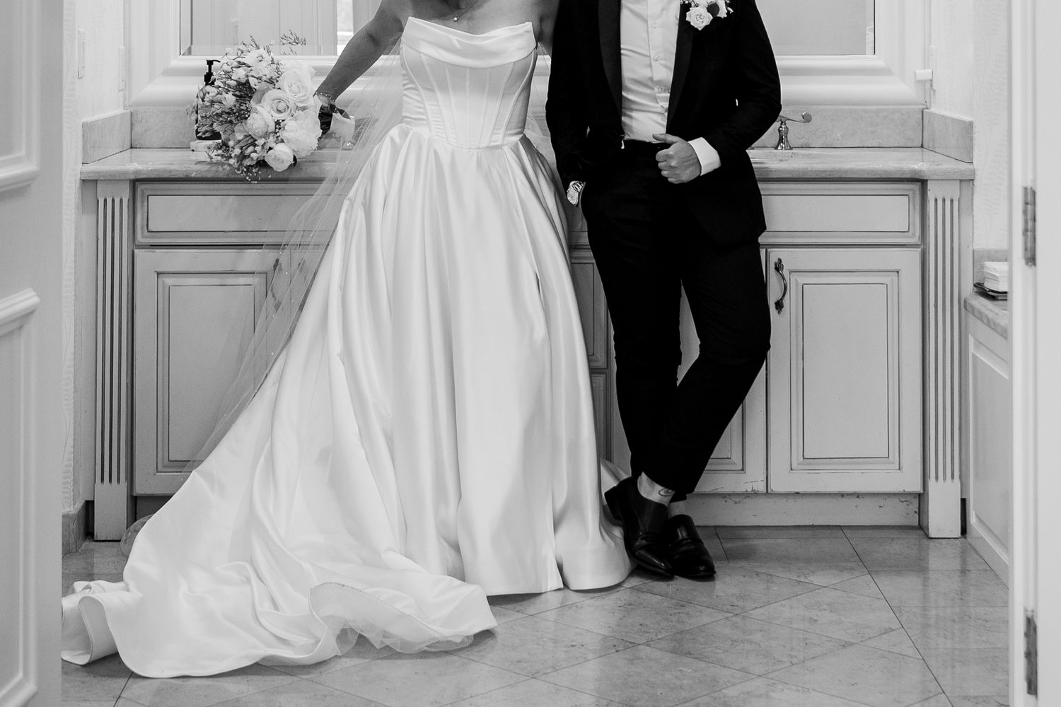 Black and white photo of a bride in a white gown and groom in a tuxedo standing together near elegant cabinetry.