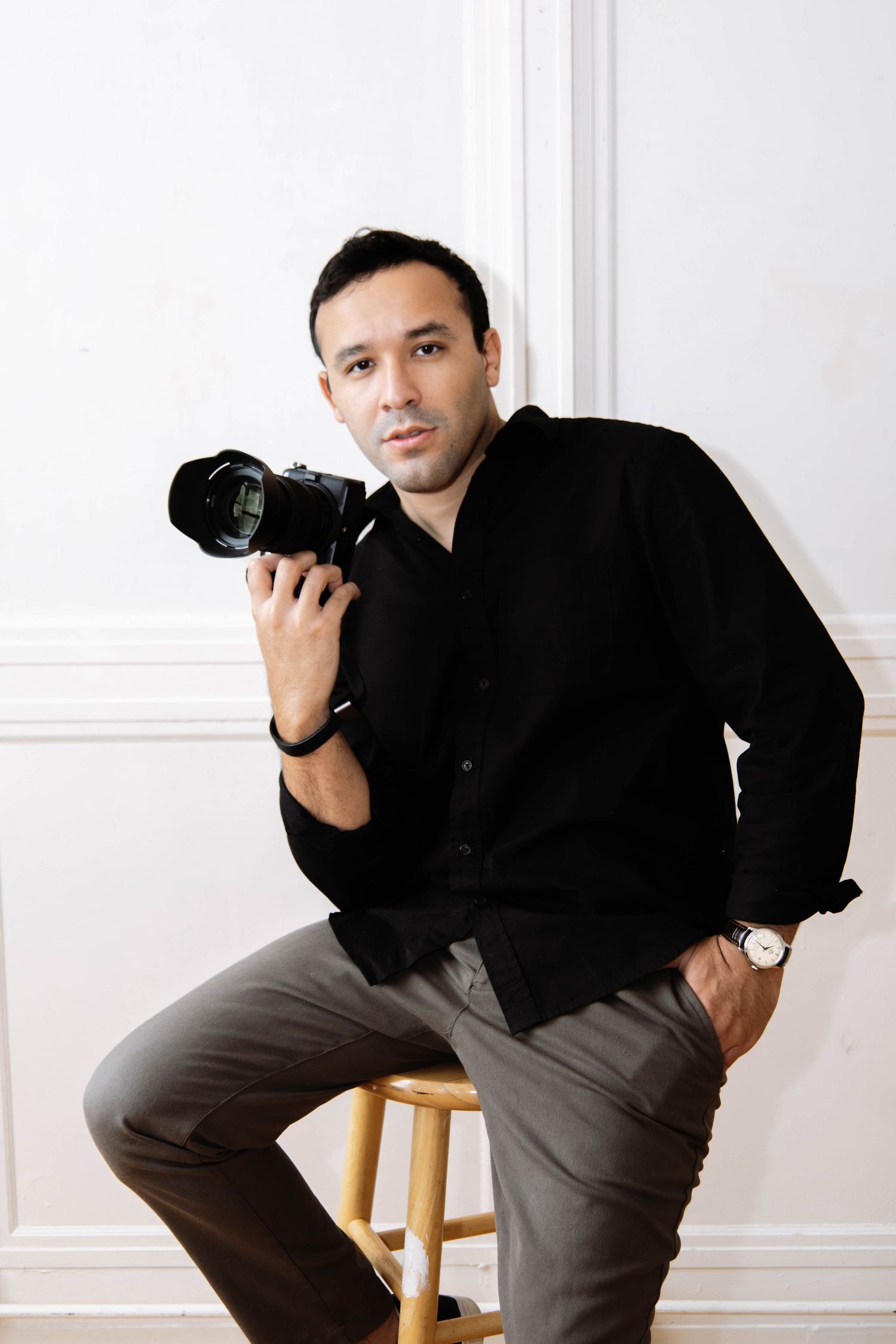 Professional photographer in black attire sits on wooden stool while posing with camera in studio setting.