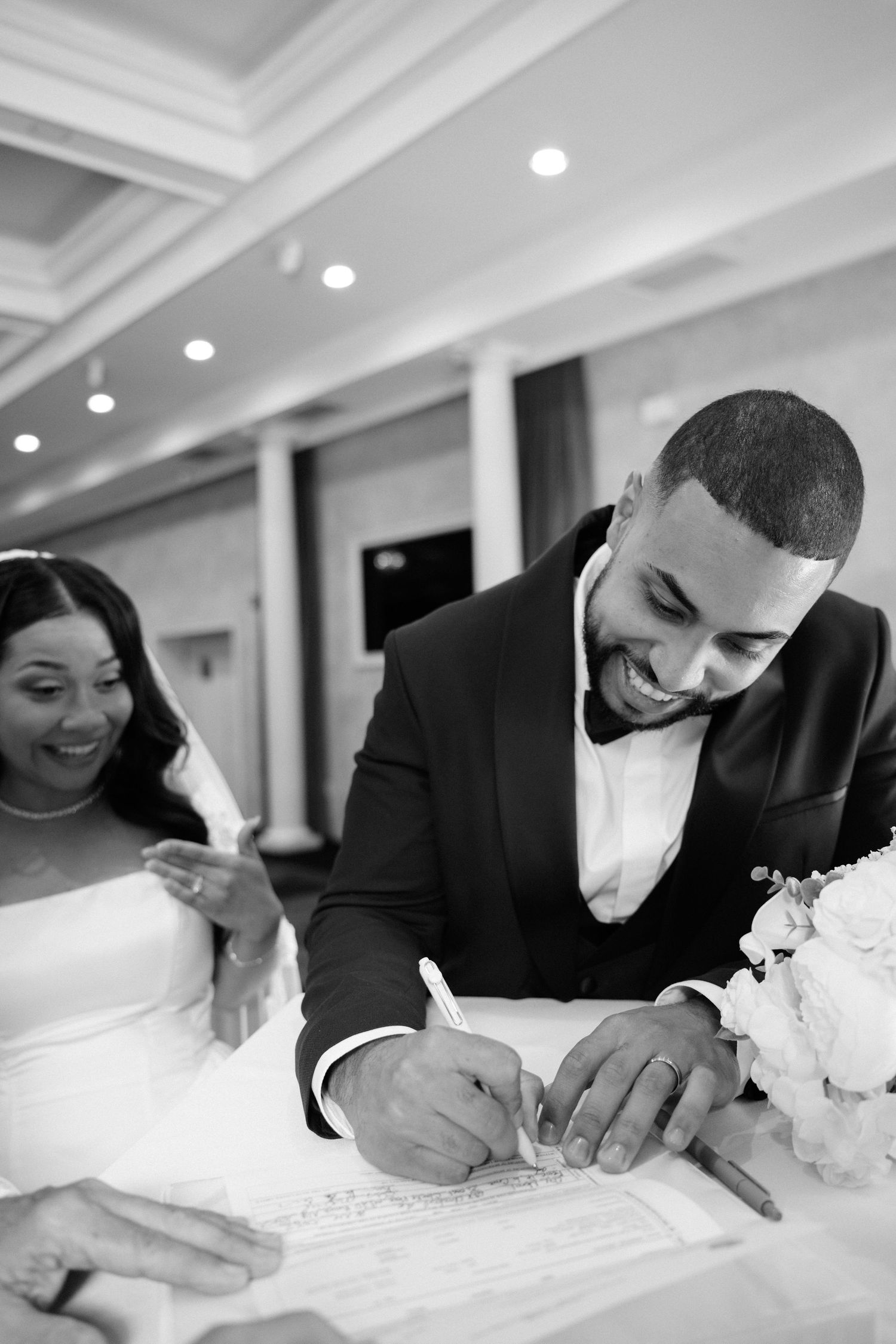 Black and white photo of a joyful groom signing wedding documents beside his smiling bride in a elegant venue.