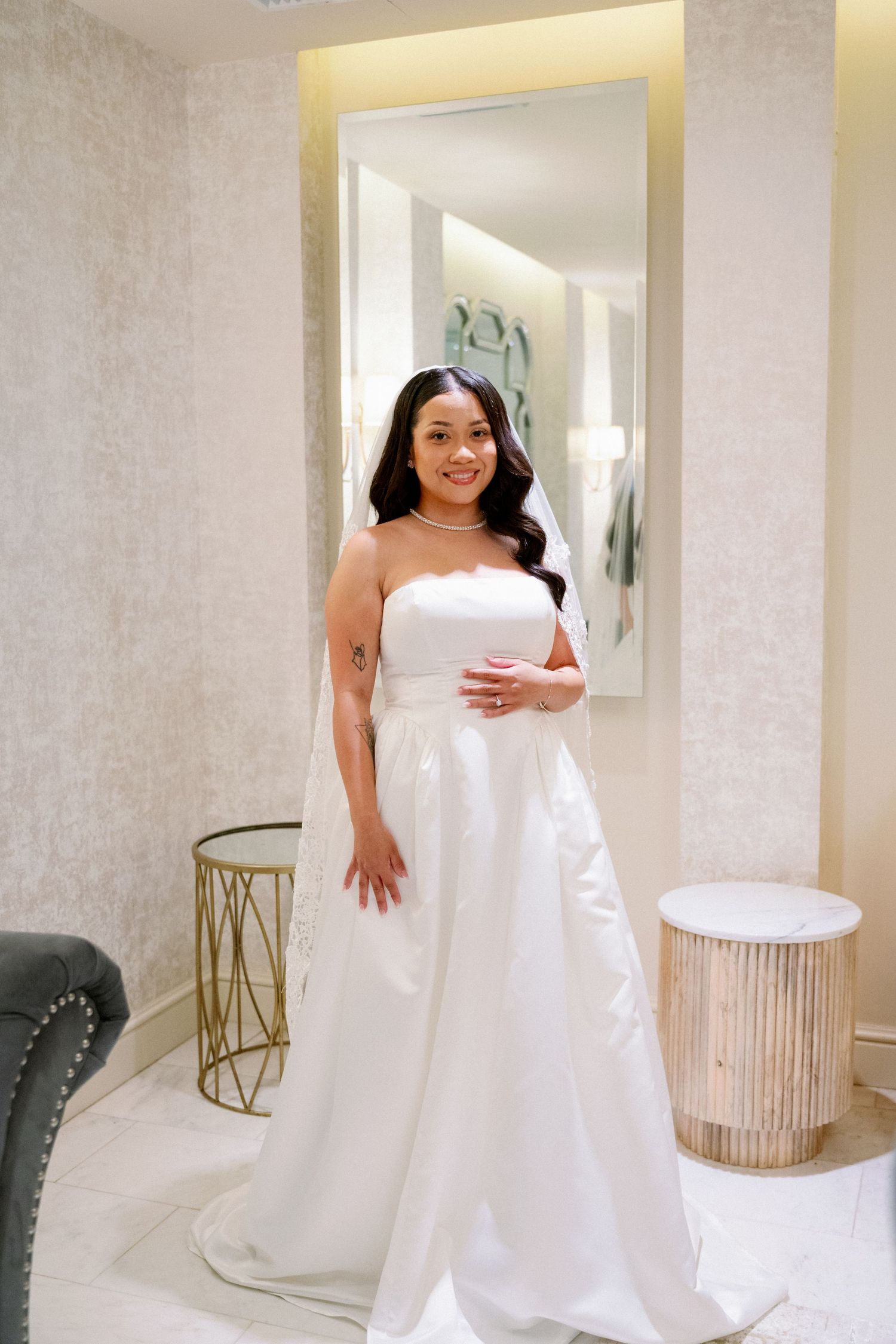 A smiling bride in a strapless white wedding gown with a veil poses in an elegant bridal boutique fitting room.