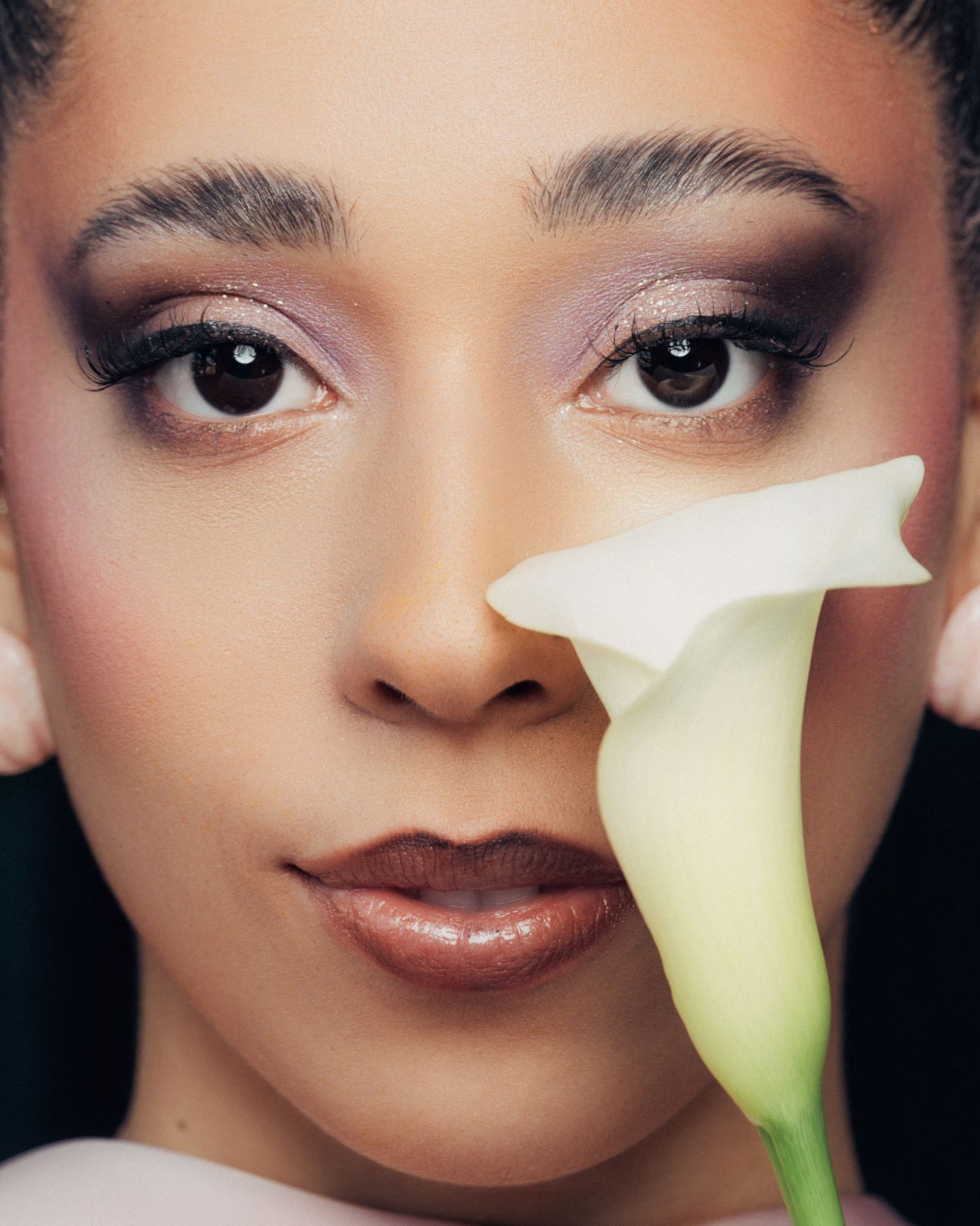 Close-up portrait of a woman with smoky purple eye makeup holding a white calla lily flower near her face.