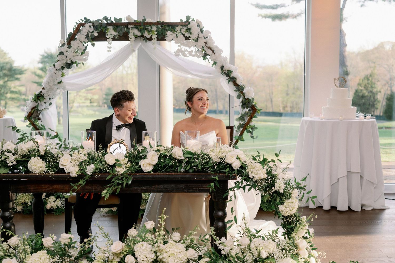 Bride and groom seated at floral sweetheart table adorned with white roses and greenery arch in elegant venue.
