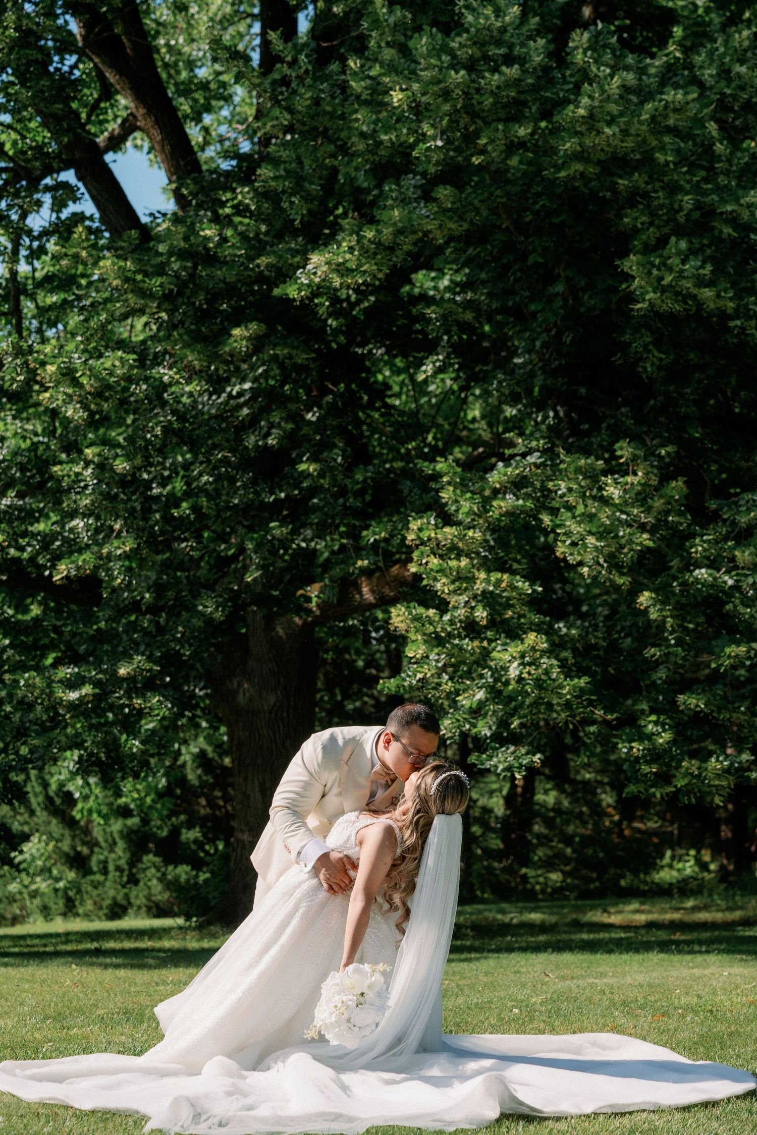 Groom in ivory suit dips bride in white gown for romantic kiss outdoors in lush green park setting.