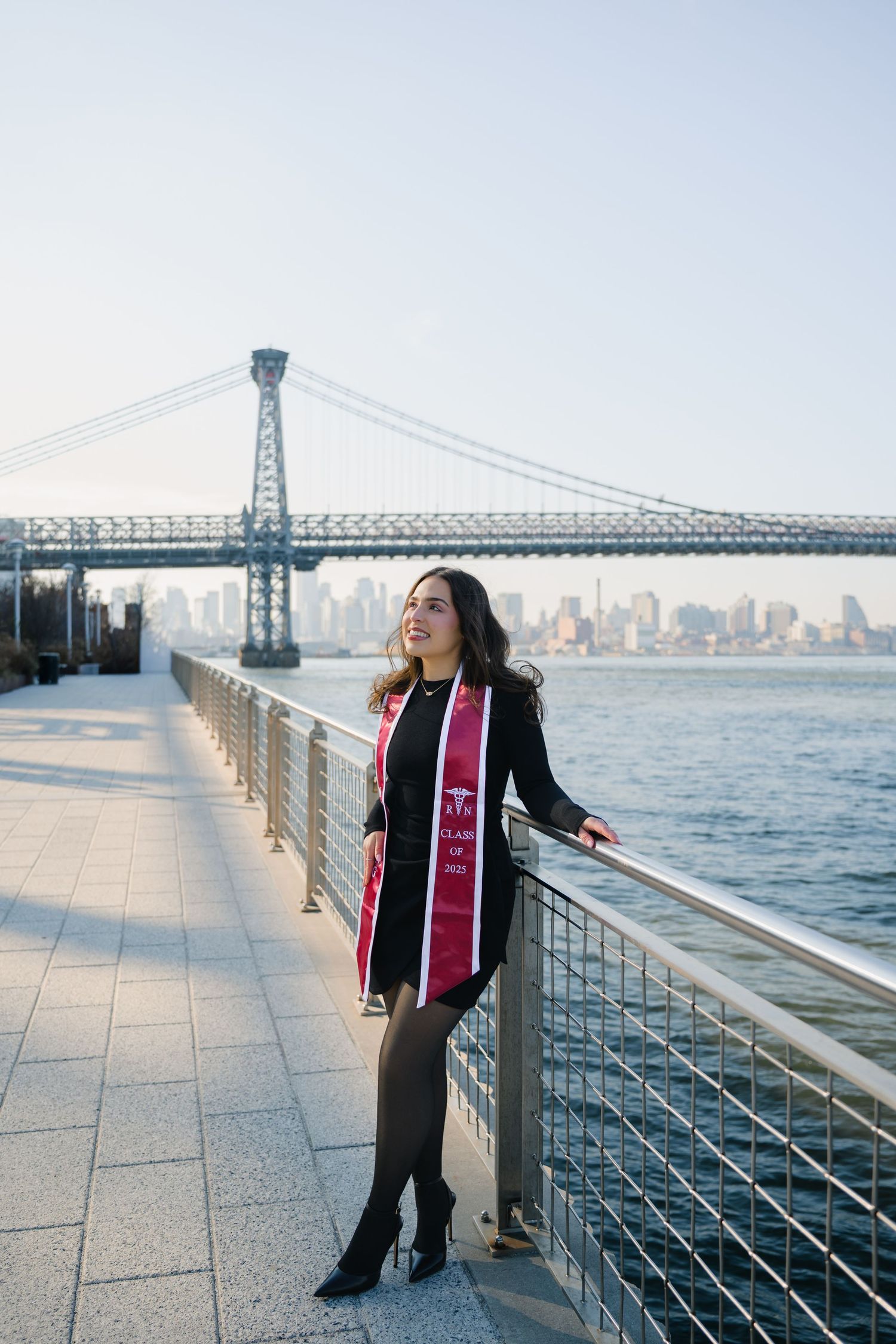 A graduate poses for photos along the waterfront with the Williamsburg Bridge and Manhattan skyline in the background.
