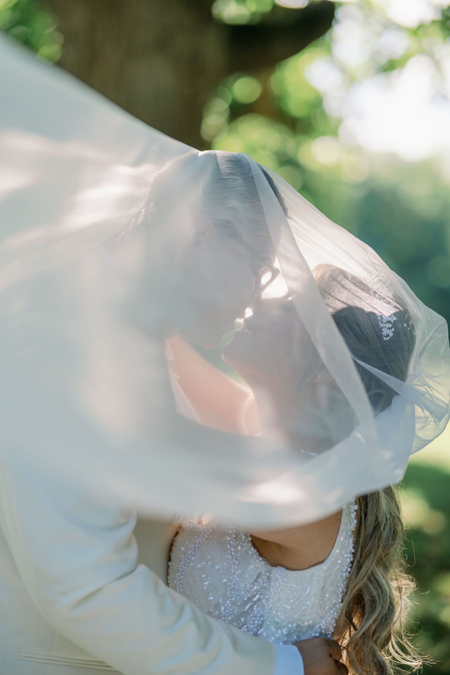 Bride and groom sharing a romantic kiss under a flowing white veil outdoors with soft green bokeh background.