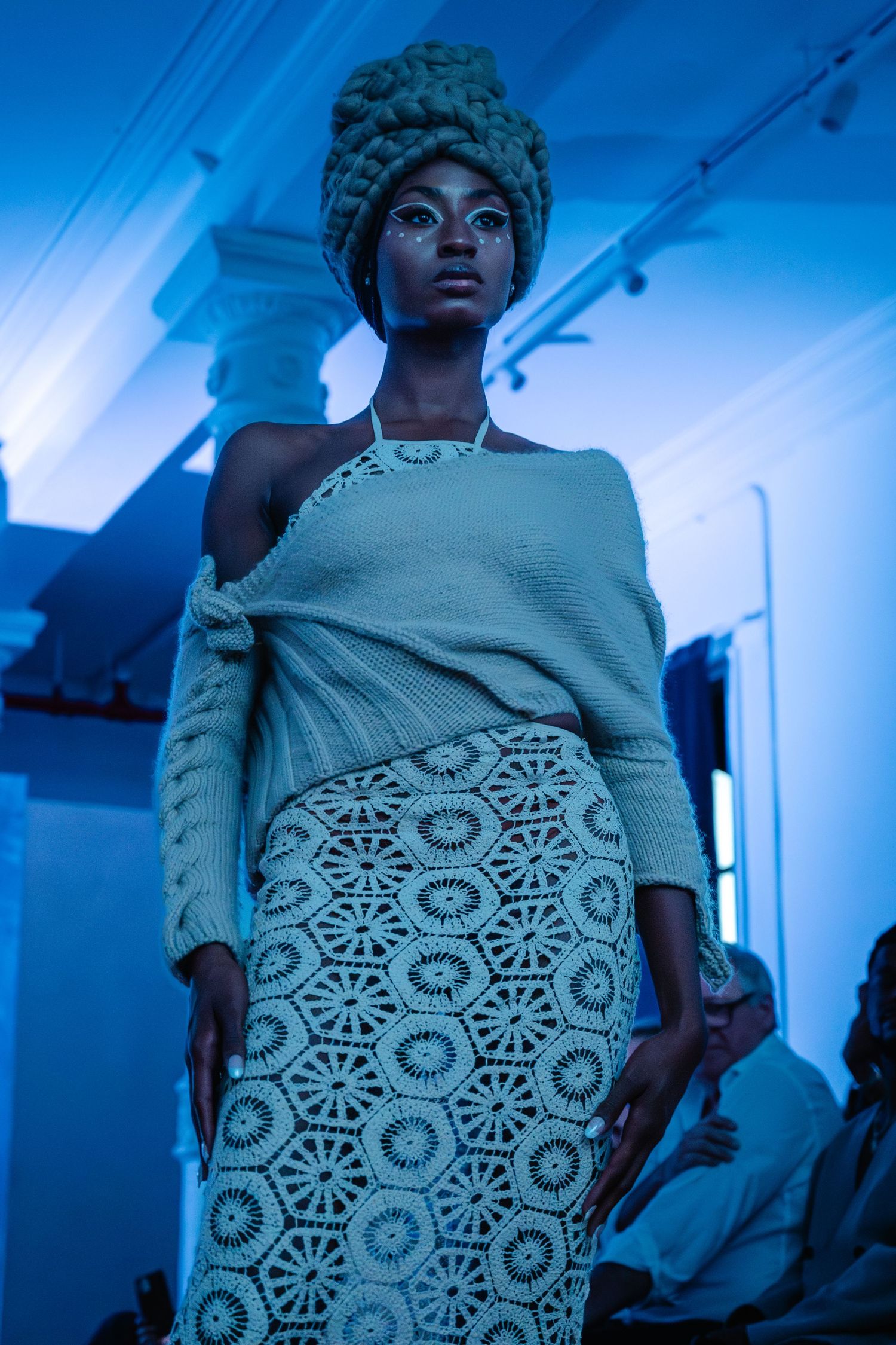 Runway model in a sage green off-shoulder knit sweater, geometric crochet skirt and matching textured turban under blue lighting.