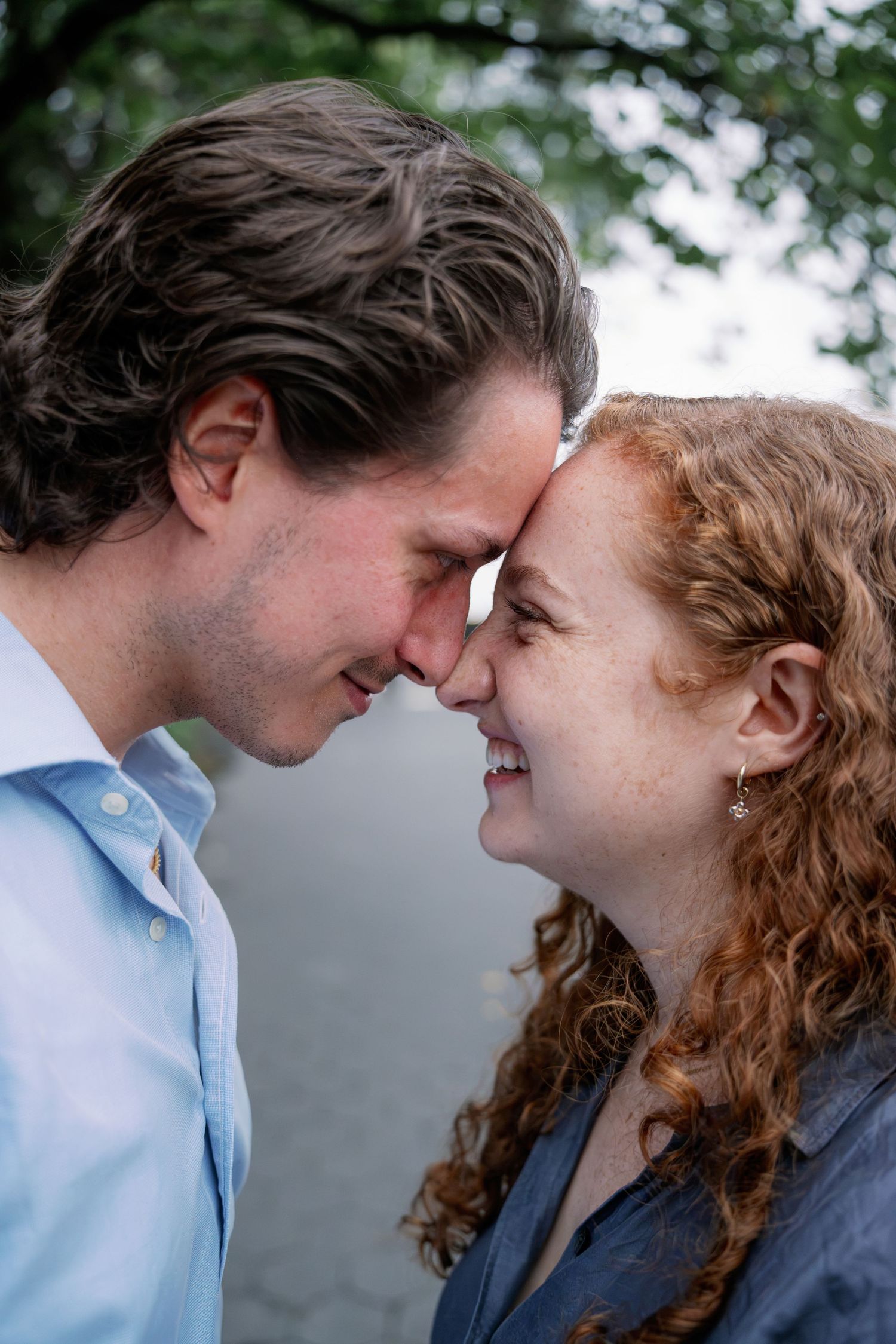 Smiling couple with foreheads touching in a tender engagement photo outdoors, man in light blue shirt and woman with curly red hair.