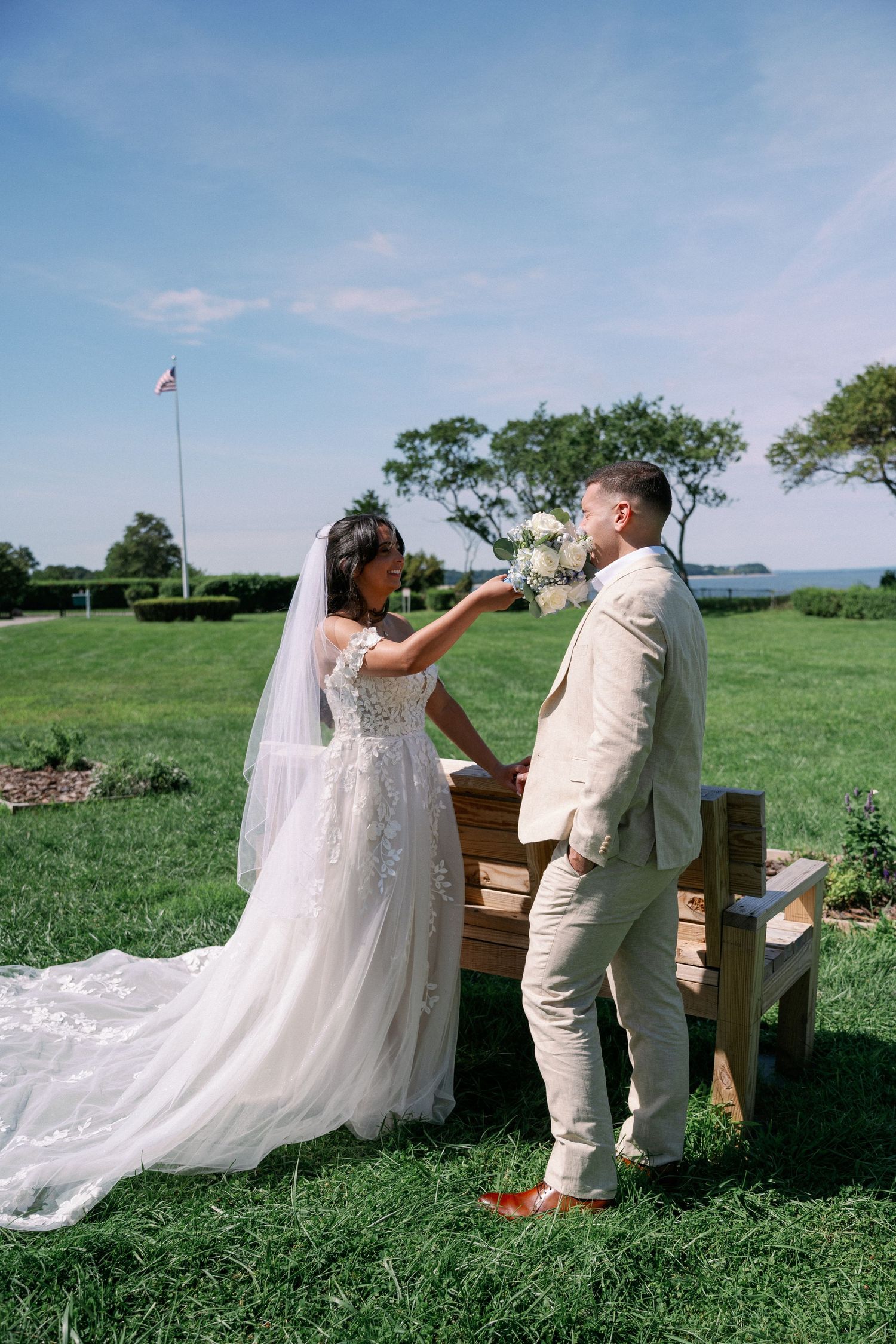 Bride in a lace gown with a long veil and groom in a cream suit share a first look beside a wooden bench on a sunny waterfront lawn.