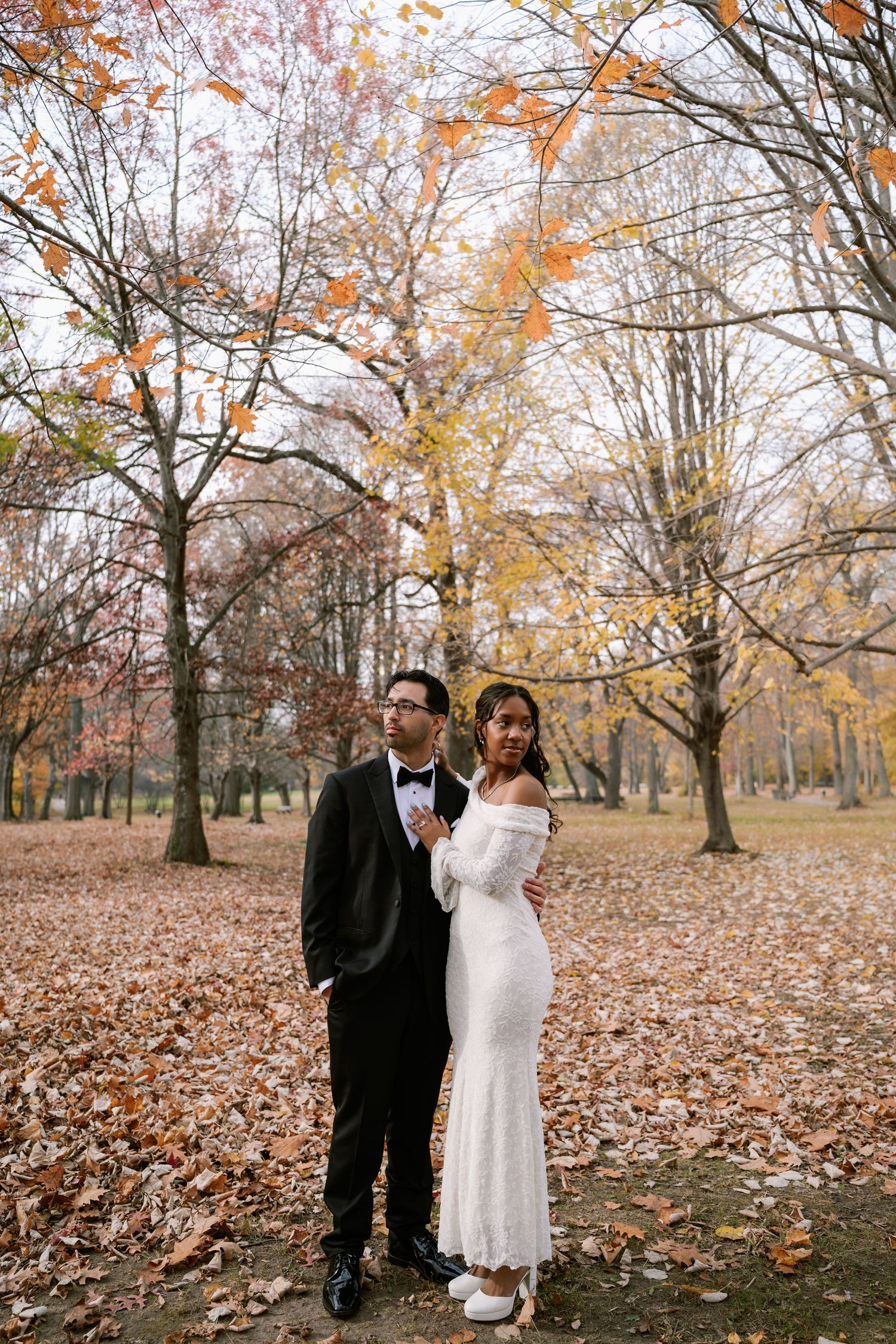A couple in formal wedding attire stands together in a park during autumn with fallen leaves on the ground.