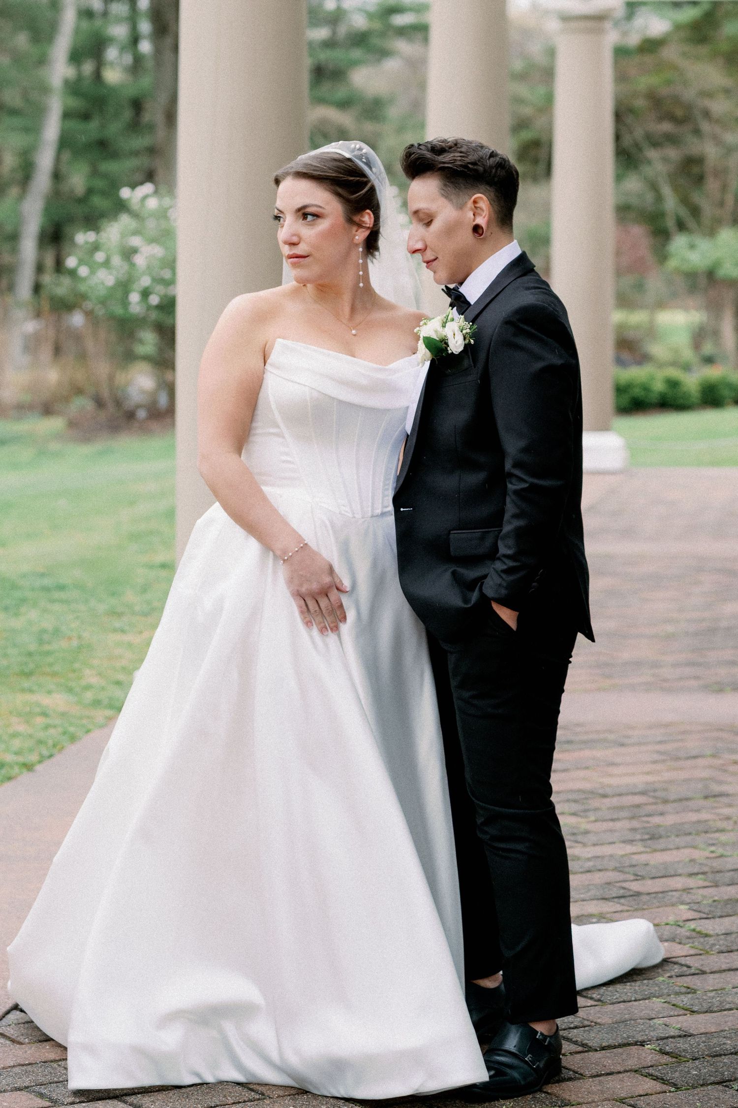 Bride in strapless white gown and groom in black tuxedo pose together on brick path with columns and greenery.