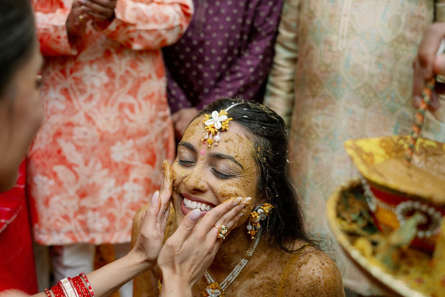 Bride smiling joyfully during a traditional Indian Haldi ceremony, with turmeric paste applied to her face.