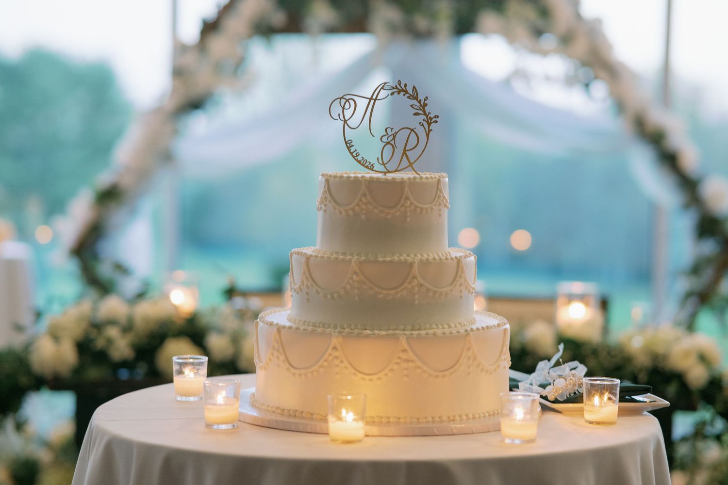 Three-tier white wedding cake with monogram topper, surrounded by candles and floral arch backdrop.