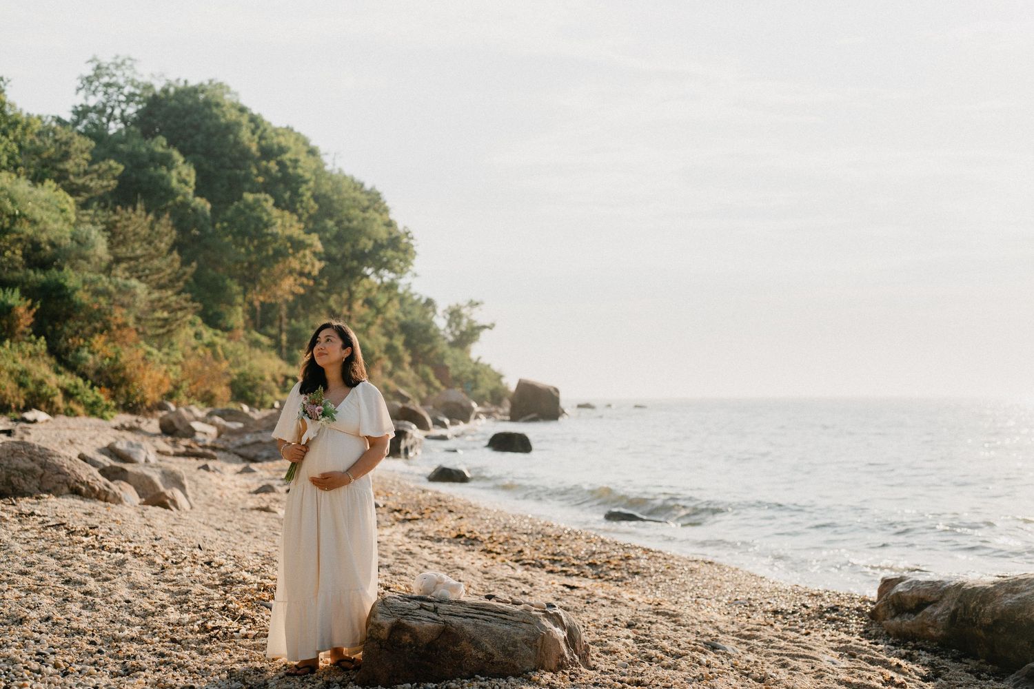 Pregnant woman in white dress standing on rocky beach holding flowers with ocean and trees in background.