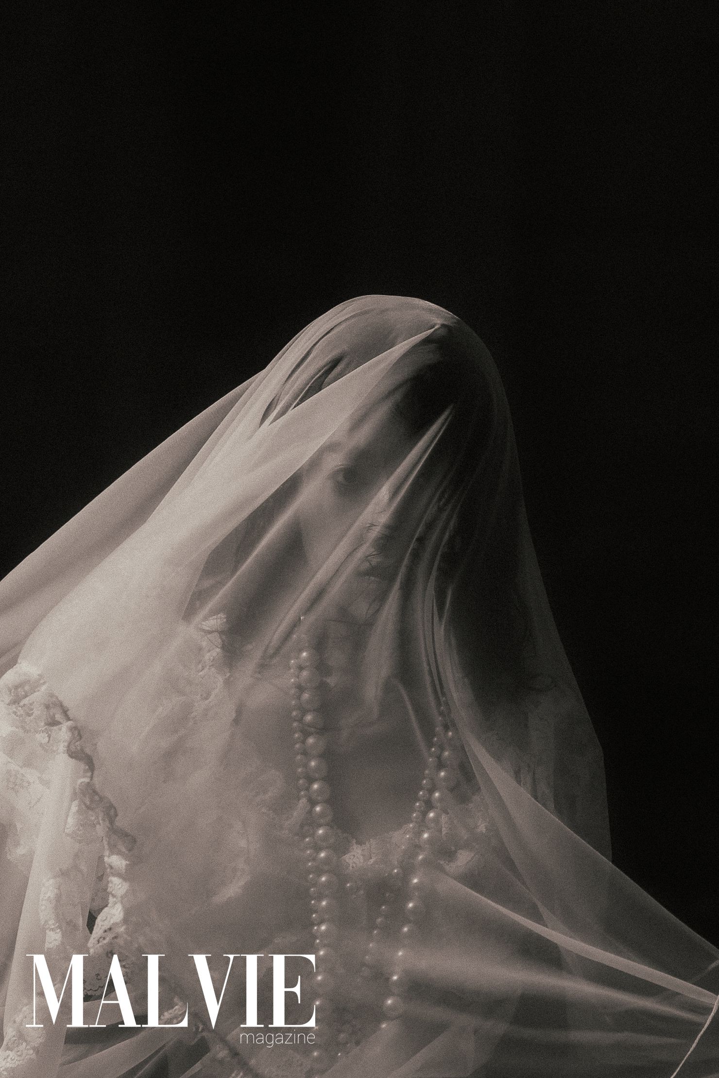 Ethereal figure draped in sheer white bridal veil with pearl necklace against dark background, Malvie Magazine editorial.