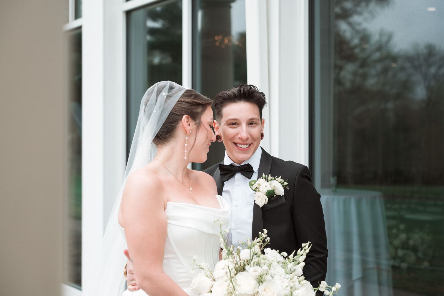 Same-sex couple on wedding day, bride in white gown with veil holding bouquet, partner in black tuxedo smiling outdoors.