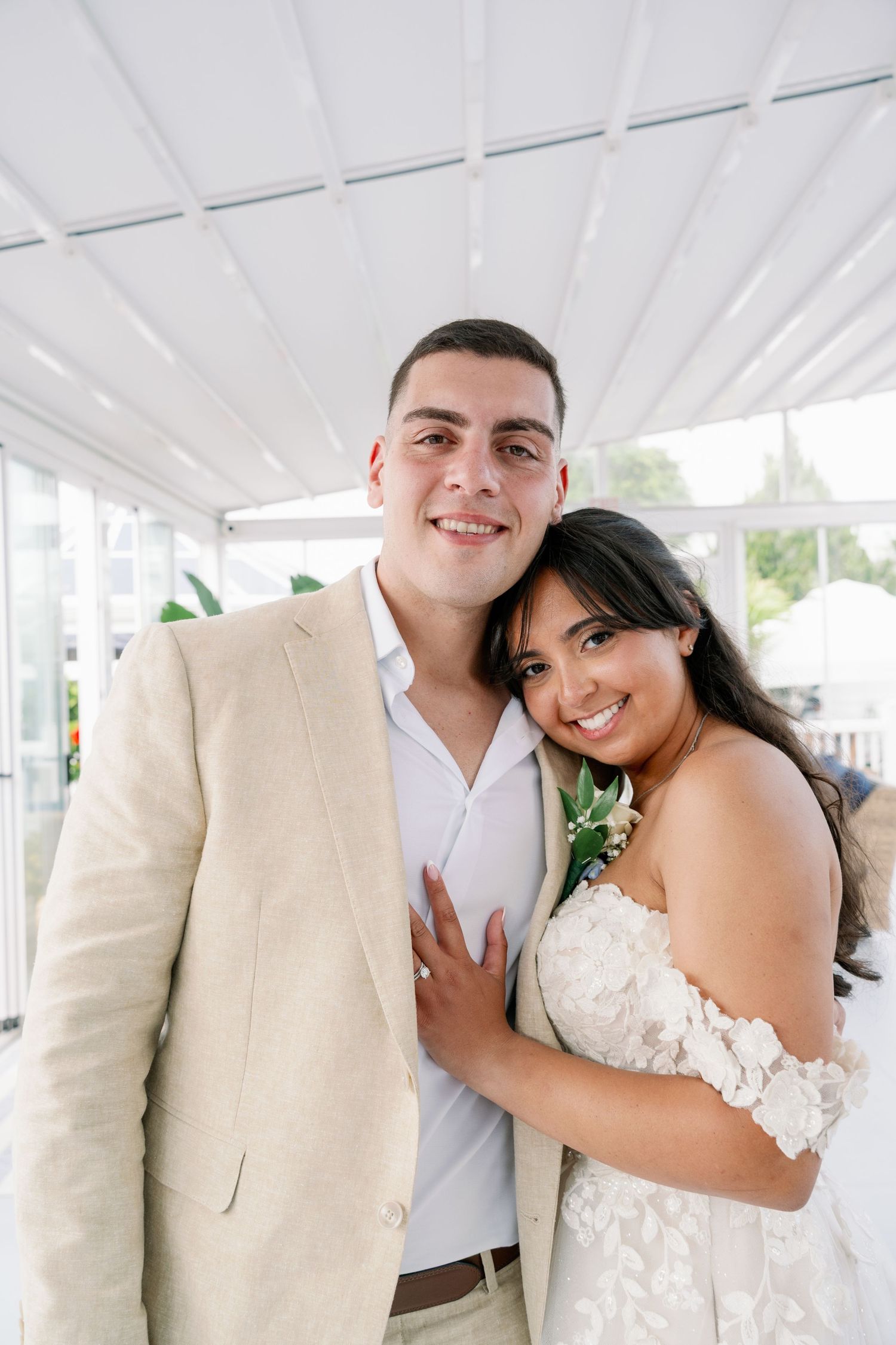 Smiling bride in a white lace off-the-shoulder wedding dress embracing a groom in a beige blazer inside a bright, airy venue.