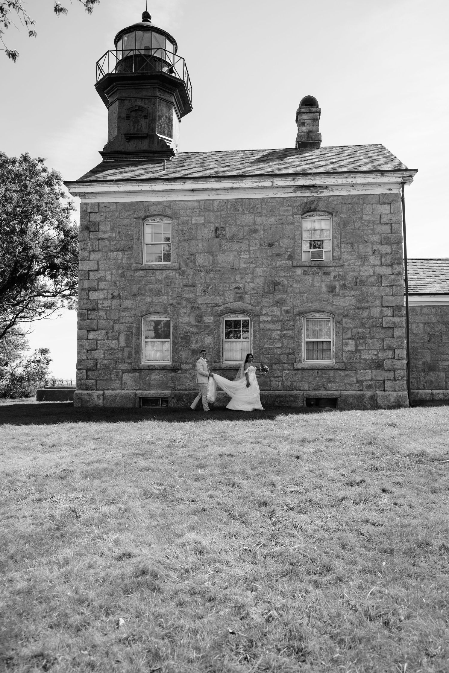Black and white photo of a couple in wedding attire walking on the lawn in front of a historic stone lighthouse building.