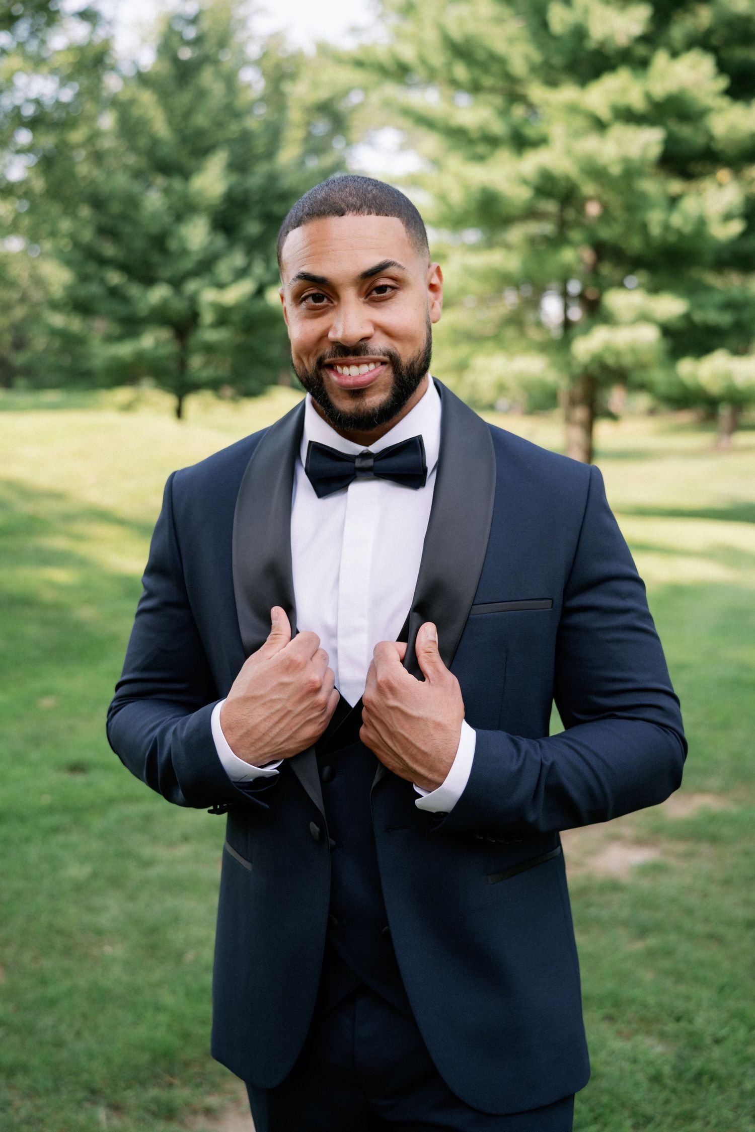 A smiling groom in a navy tuxedo with black bow tie poses outdoors on green grass with trees in the background.